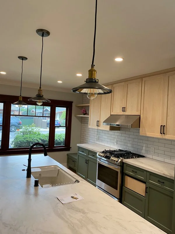Modern kitchen with a white marble island, black faucet, wooden upper cabinets, green lower cabinets, white subway tile backsplash, stainless steel stove, and hanging pendant lights. Large window shows outside view.