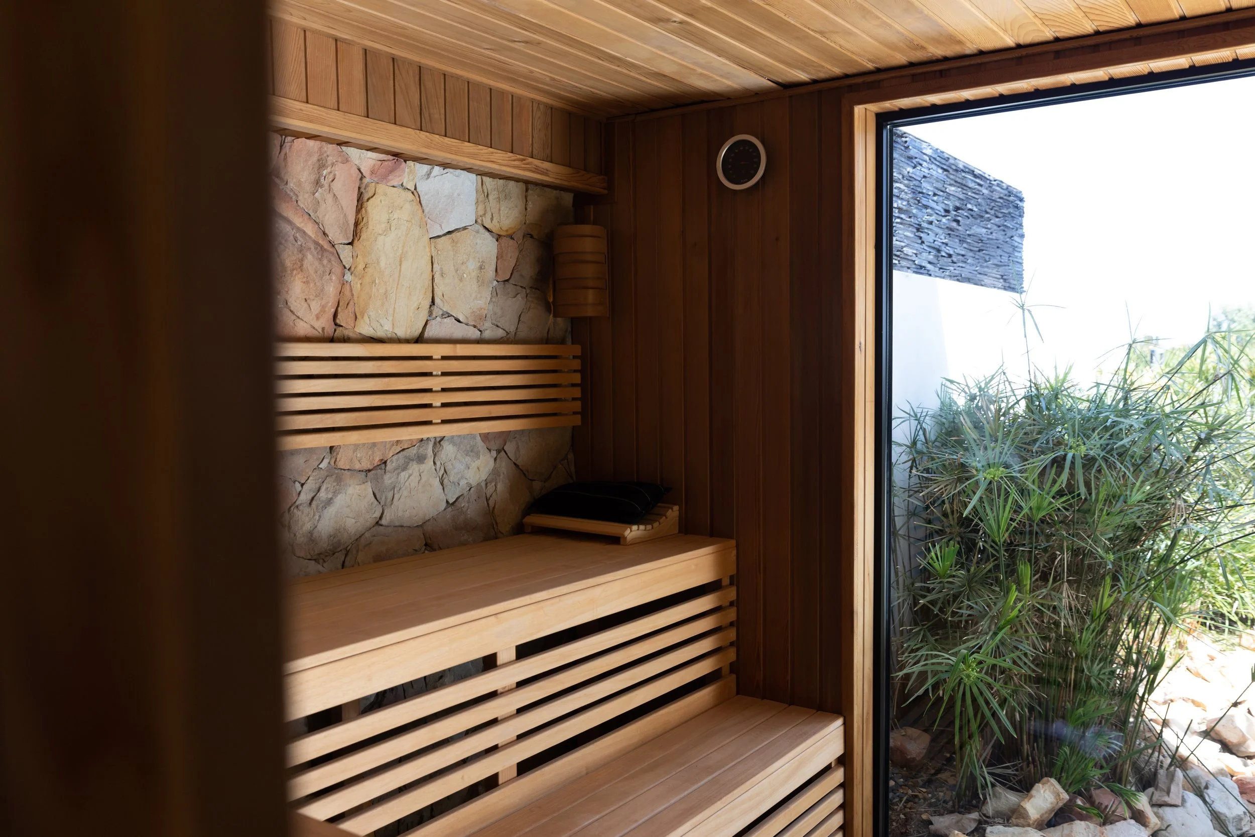 Interior of a wooden sauna with slatted benches, a stone wall, a black round thermometer, and a large window showing outdoor plants and rocks.