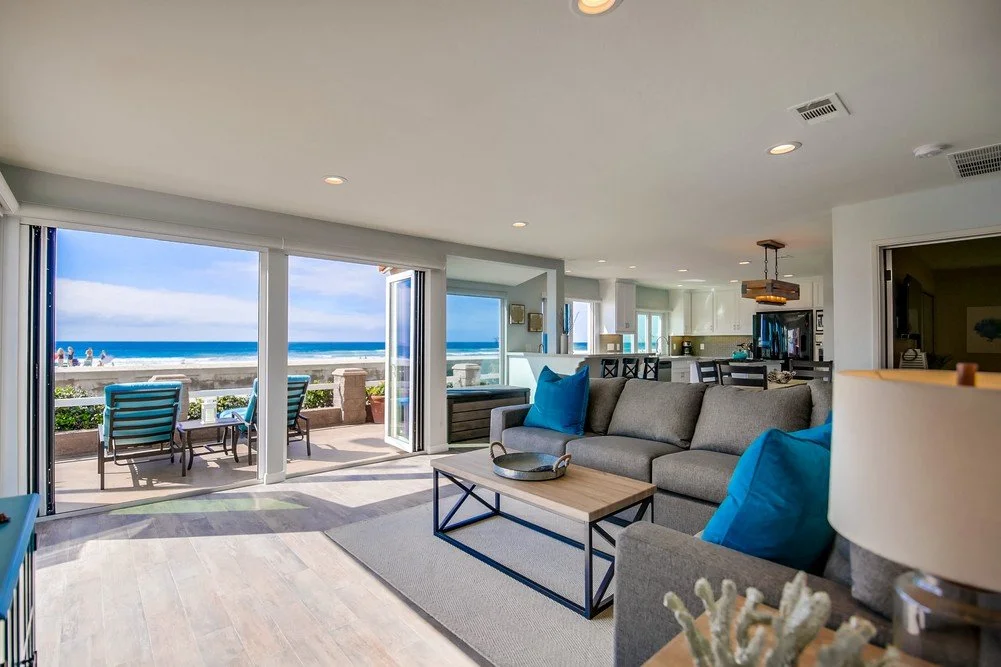 Living room with a view of the beach, featuring a gray sofa with blue pillows, a wooden coffee table, and an outdoor patio with chairs and ocean view.