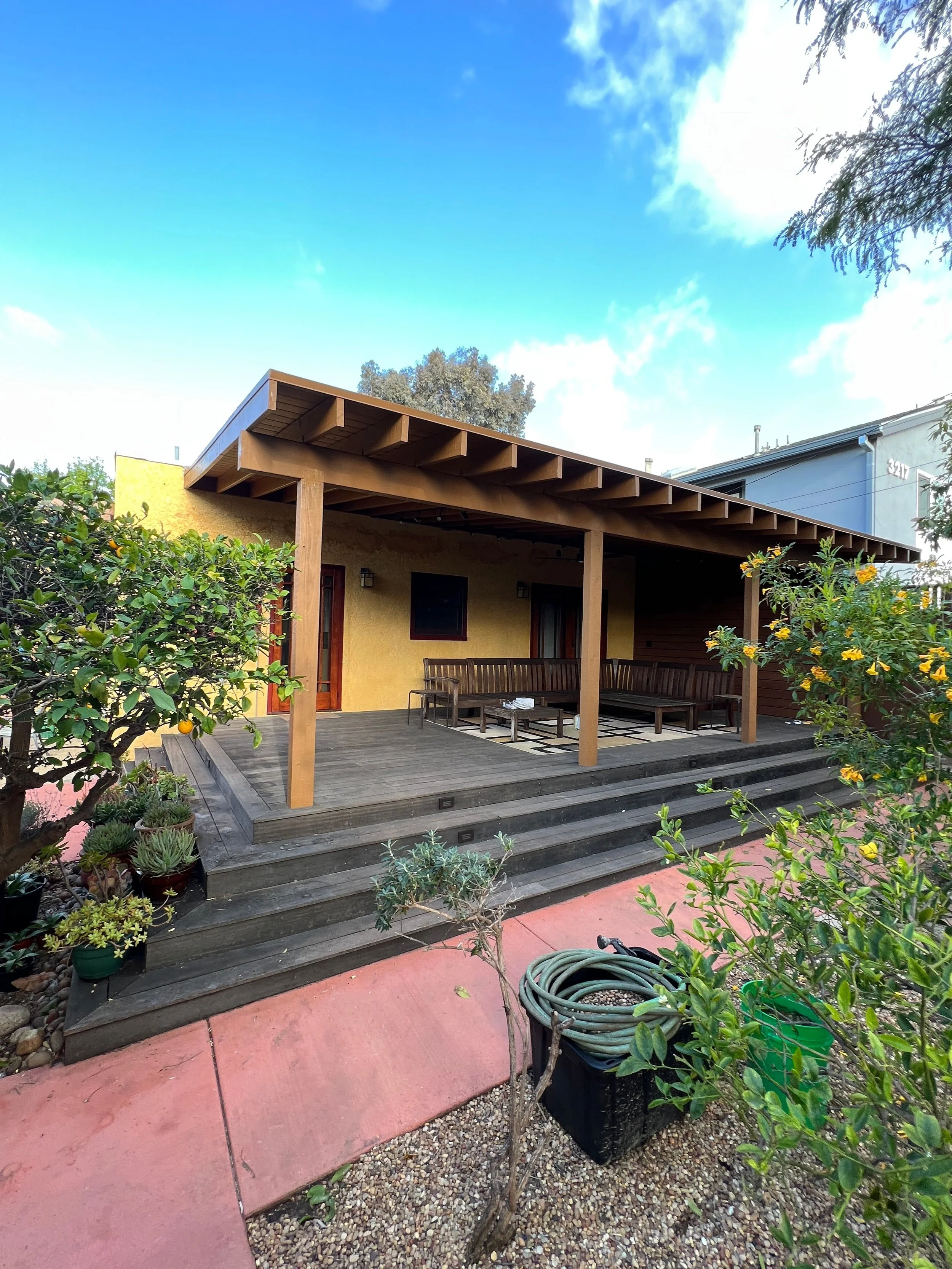 An outdoor patio area featuring wooden decking and steps leading up to a yellow house with a covered porch, surrounded by plants and trees, with a blue sky and clouds overhead.