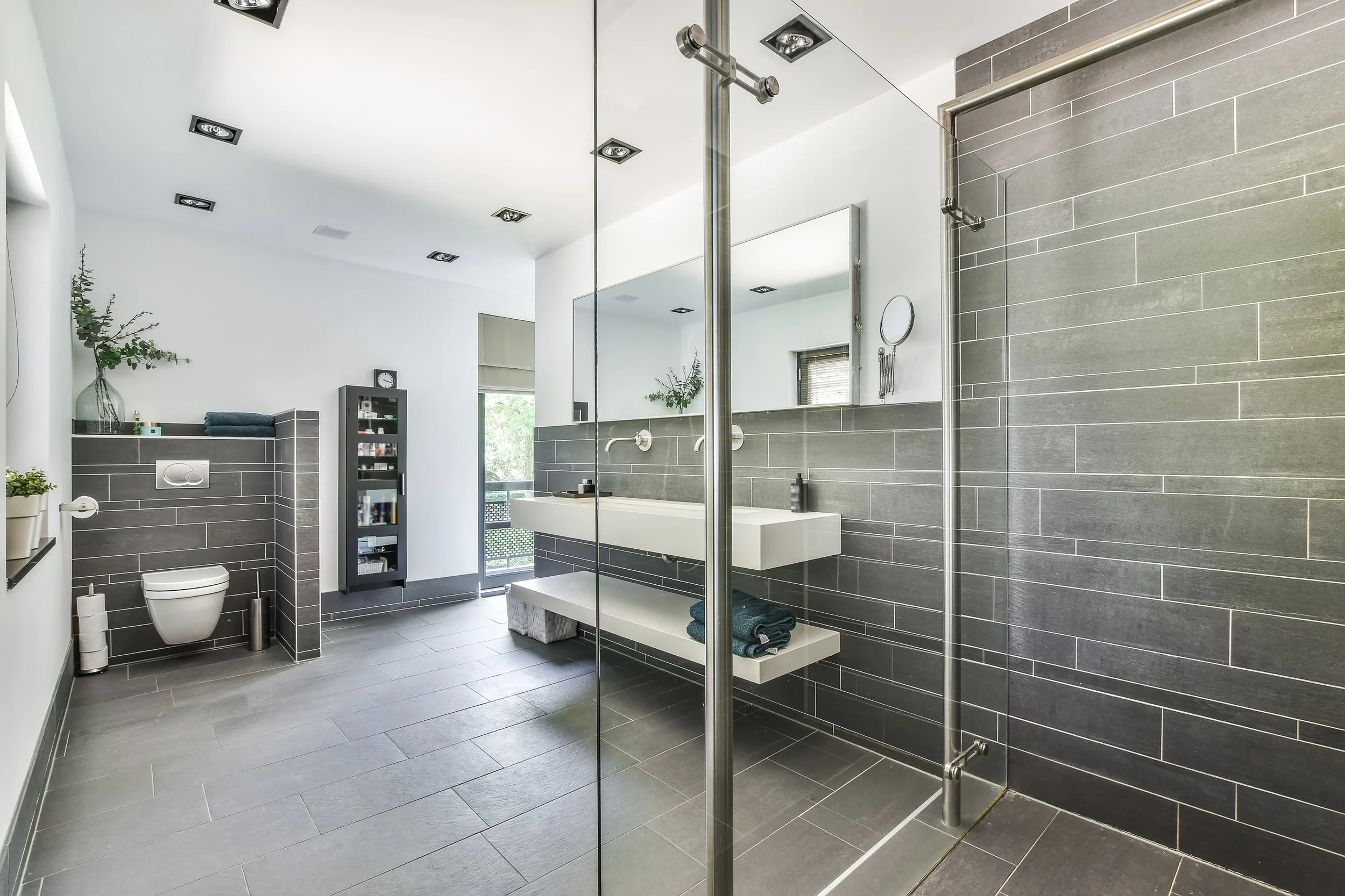 Modern bathroom with gray tile walls and floor, glass shower enclosure, white sink, wall-mounted toilet, and greenery on top of the tiled wall.