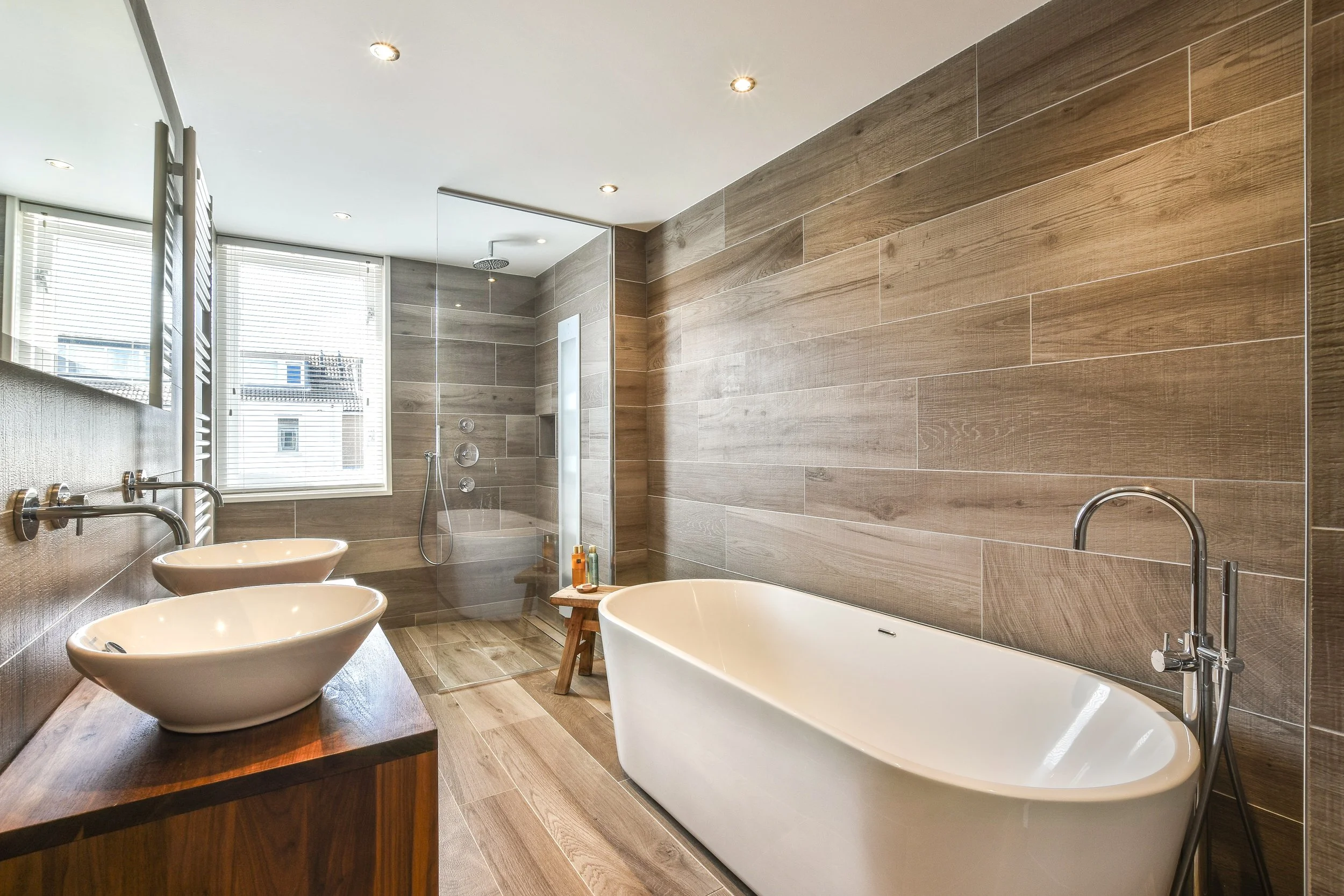 Modern bathroom with wooden wall tiles, a freestanding bathtub, and a shower with a rain shower head. There are two sinks on a wooden counter, a large window with blinds, and ceiling lights.