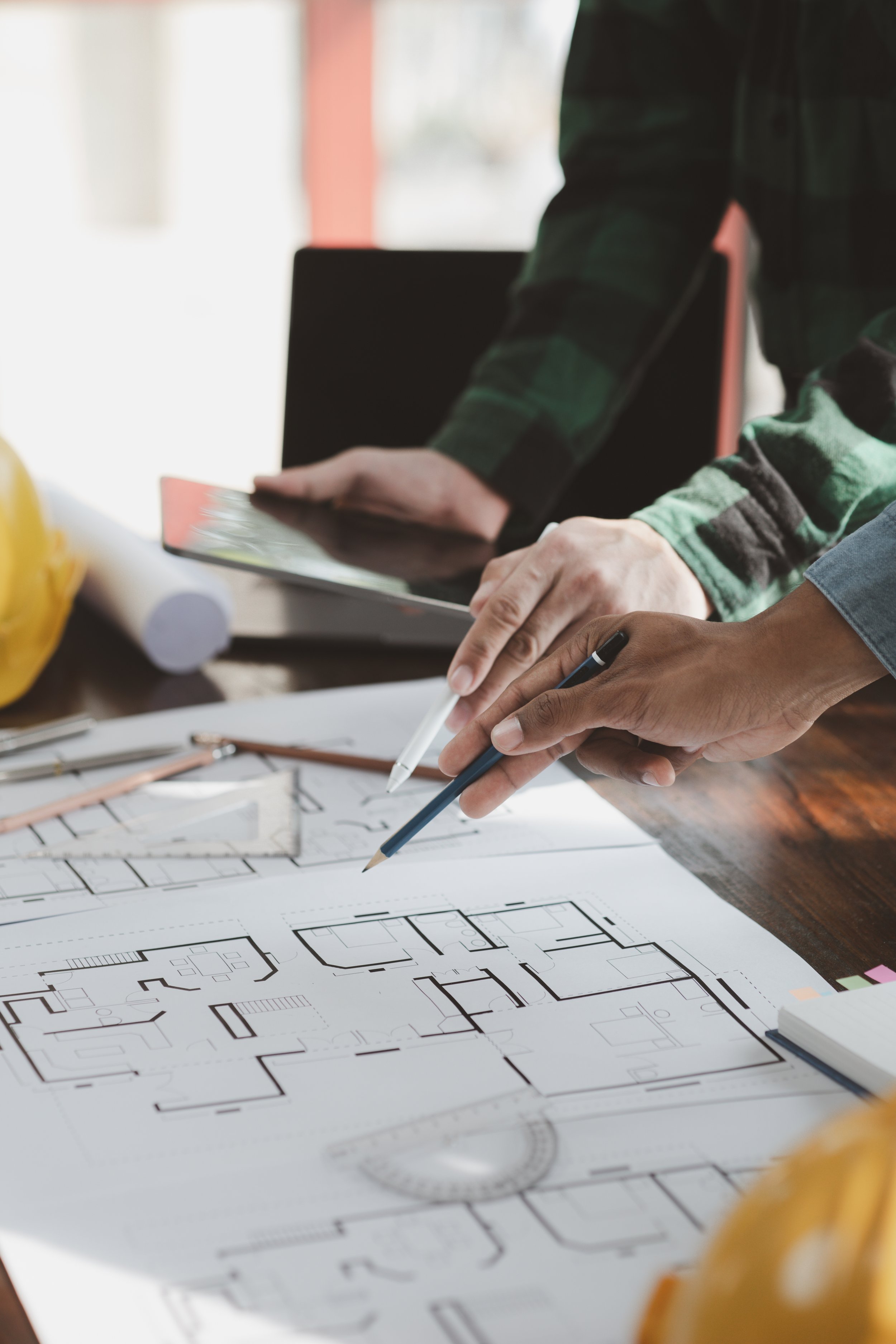 Architectural plans and blueprints on a desk with people discussing and pointing at the drawings.