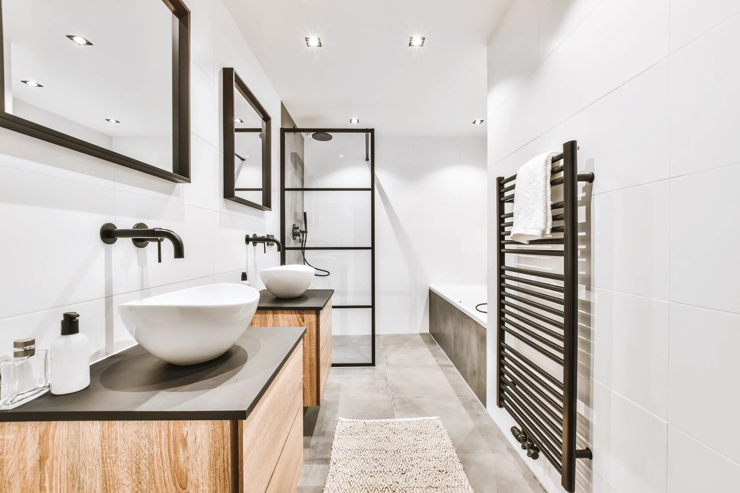 Modern bathroom with a black-framed shower enclosure, white tiled walls, a wooden vanity with a vessel sink, black faucets, a large mirror, a black towel warmer with a white towel, and gray tiled flooring.