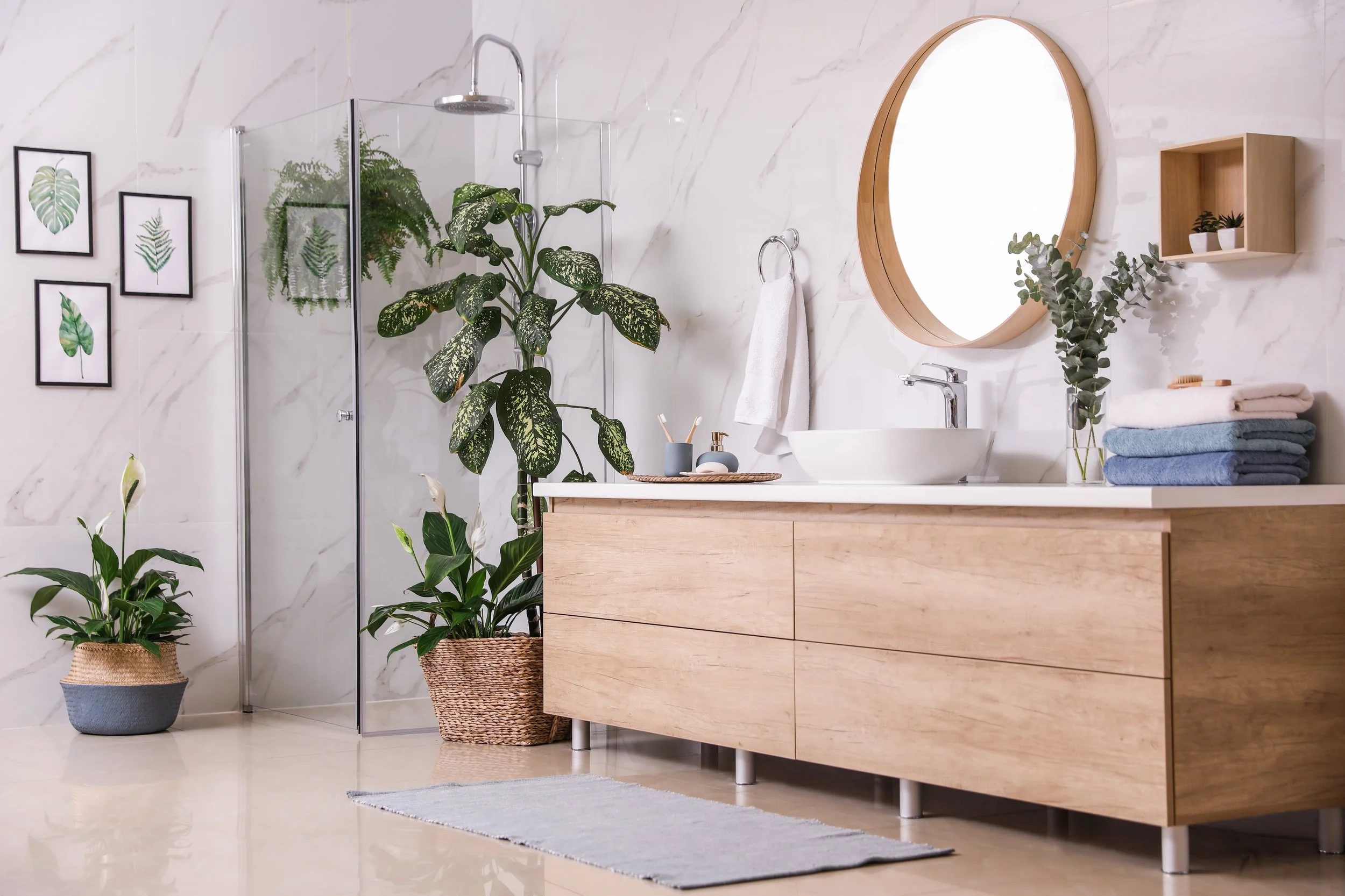 Modern bathroom with marble walls, a wooden vanity with a round mirror, plants, and towels.