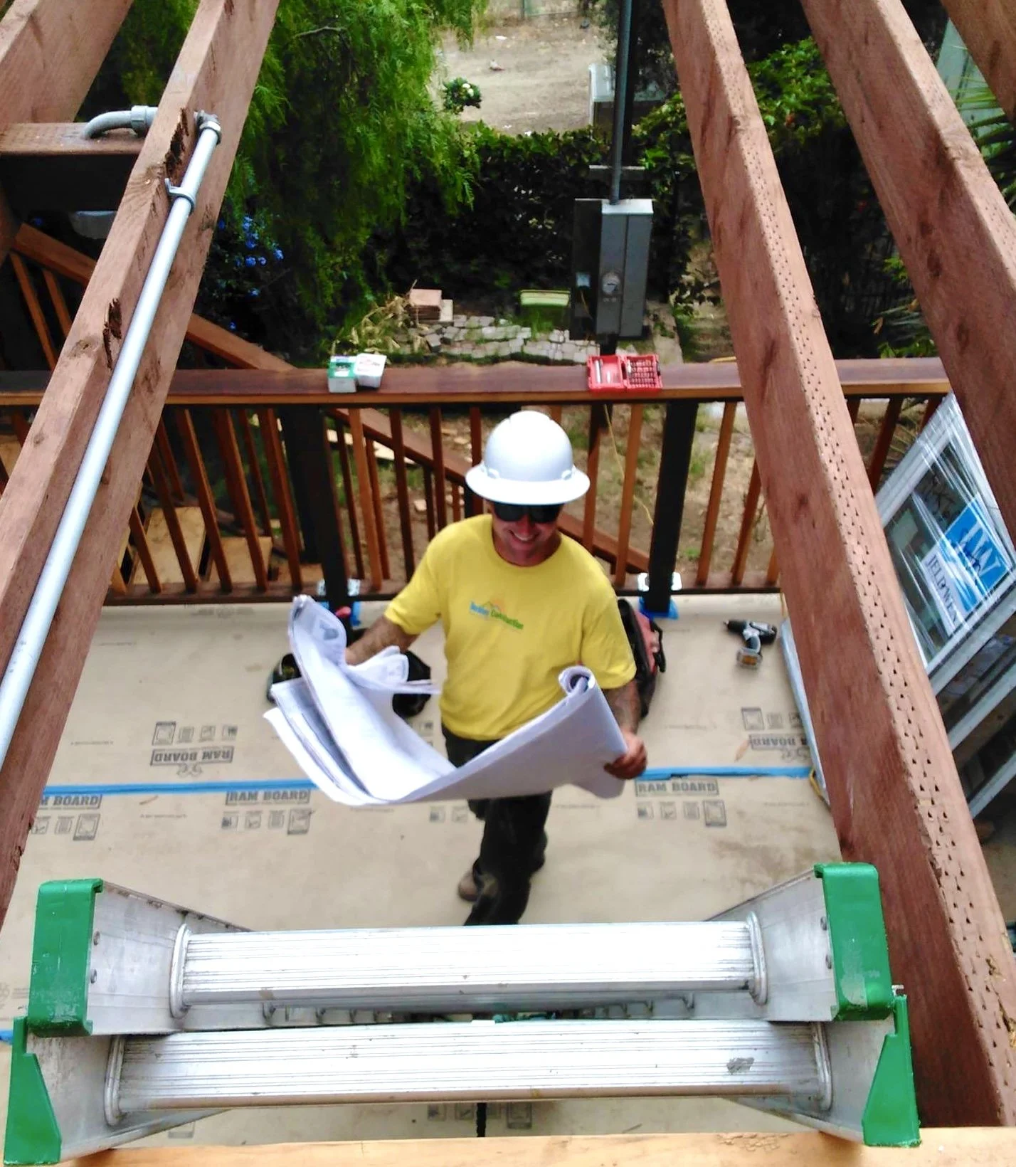 A construction worker wearing a white hard hat and yellow shirt, holding blueprints, standing inside a wooden frame of a house under construction.