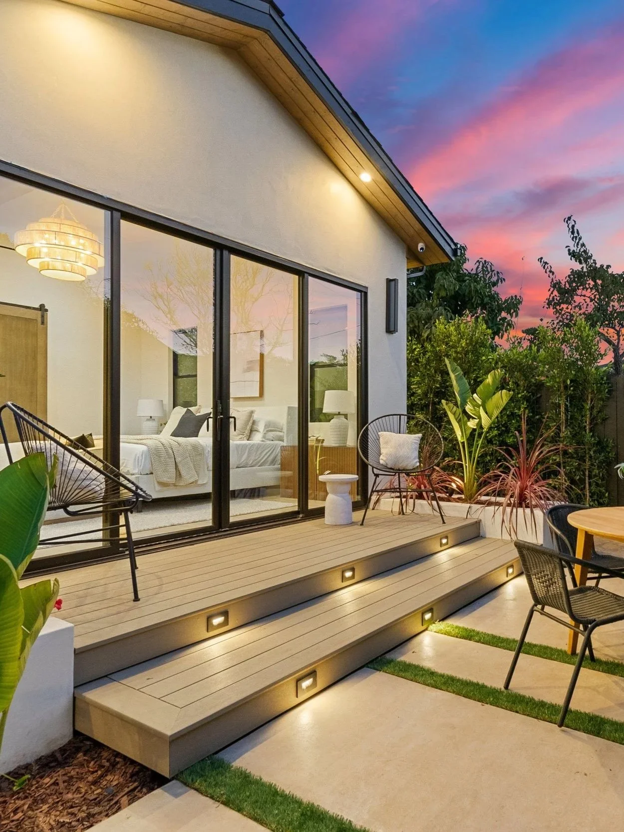 A modern house with a large glass sliding door revealing a bedroom interior, a wooden deck with outdoor seating, potted plants, and a sunset sky.