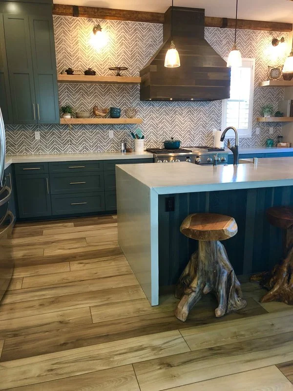 A modern kitchen with blue cabinets, open wooden shelves, a white countertop, a black range hood, and bar stools made from tree stumps.