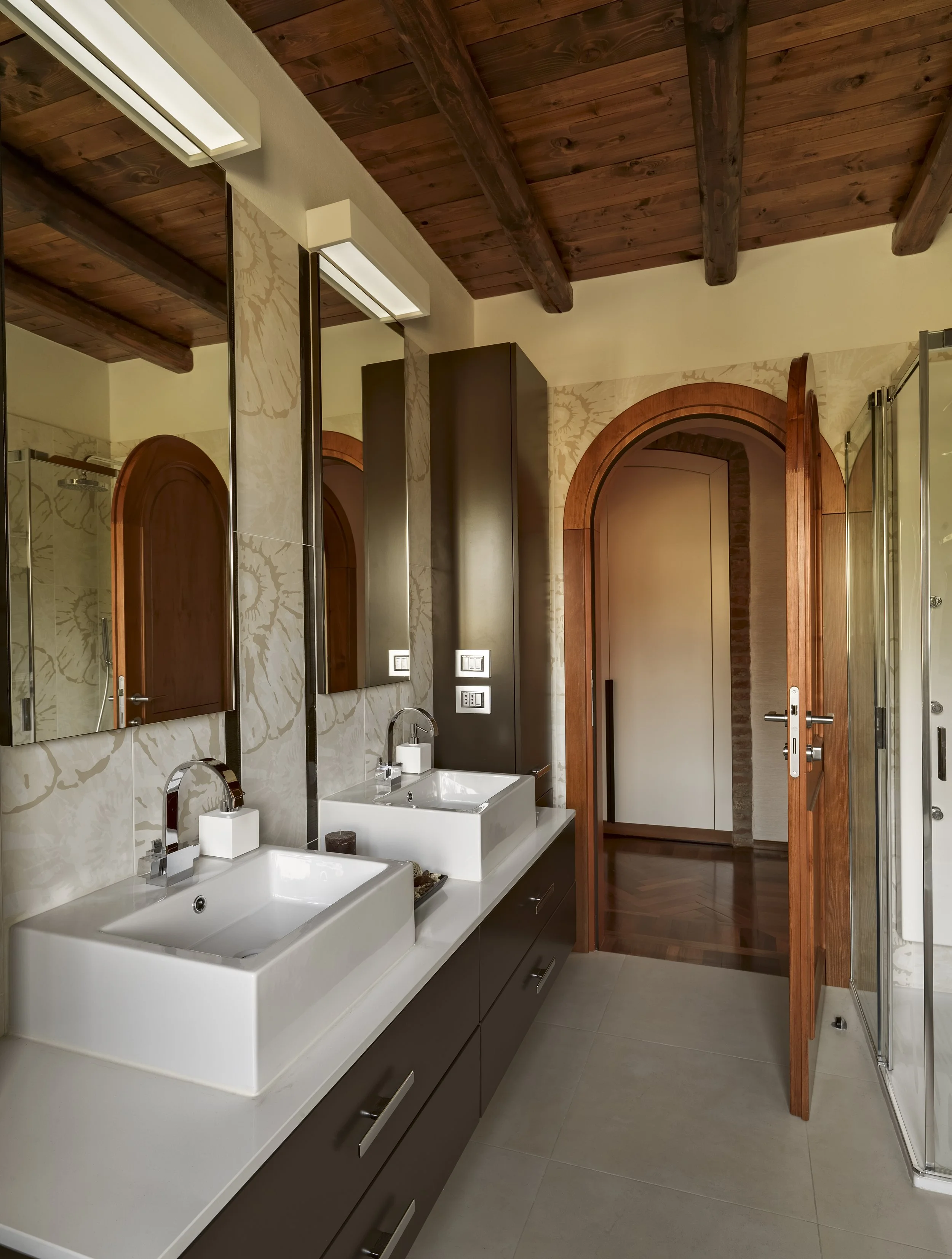 Modern bathroom with dual sinks, large mirrors, dark wood cabinetry, wood ceiling beams, and a doorway leading to another room.