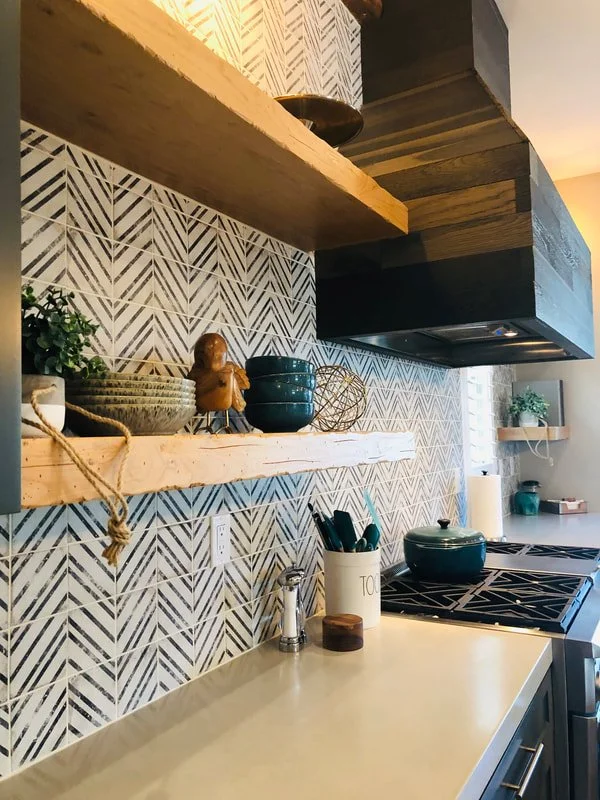 A modern kitchen with a patterned backsplash, wooden shelves holding decorative items, and teal-colored pots on the counter.
