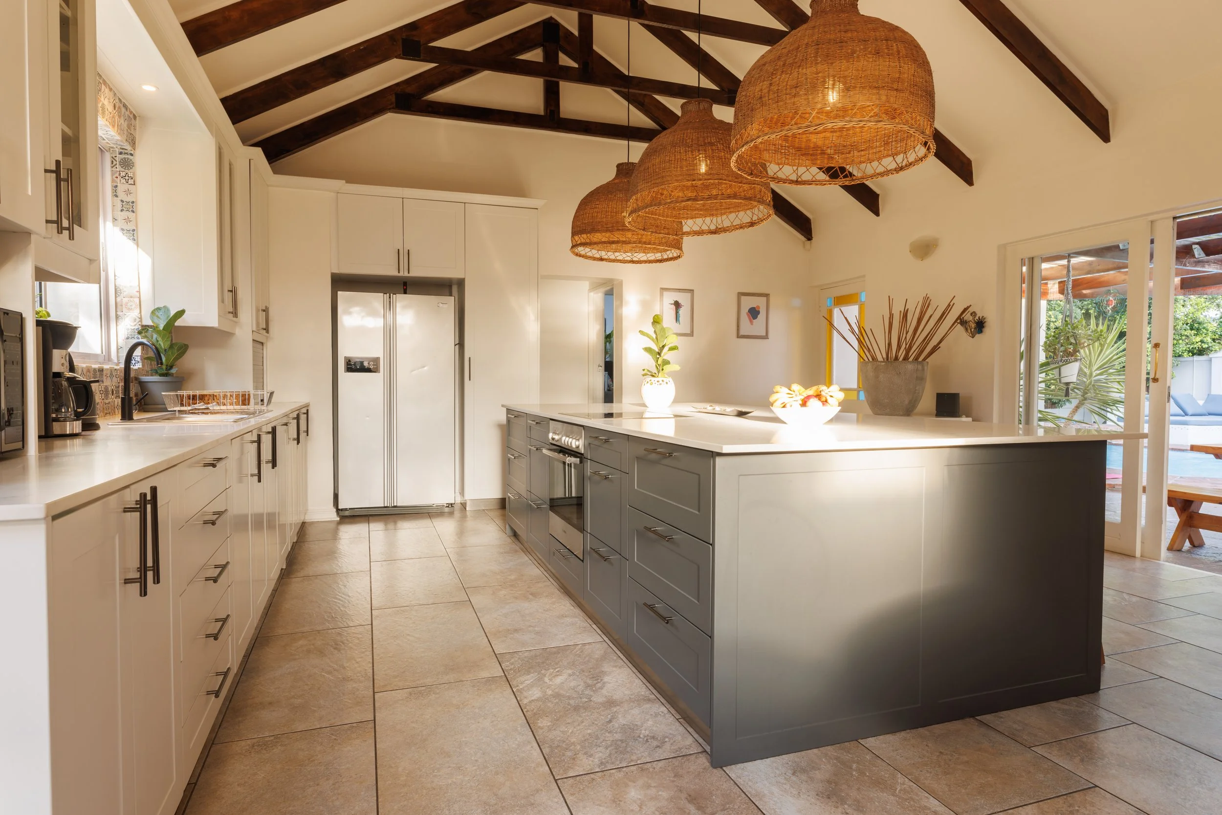 Modern kitchen with white cabinets, a gray island, hanging wicker pendant lights, light-colored tile floor, and large sliding glass door opening to a patio with greenery.