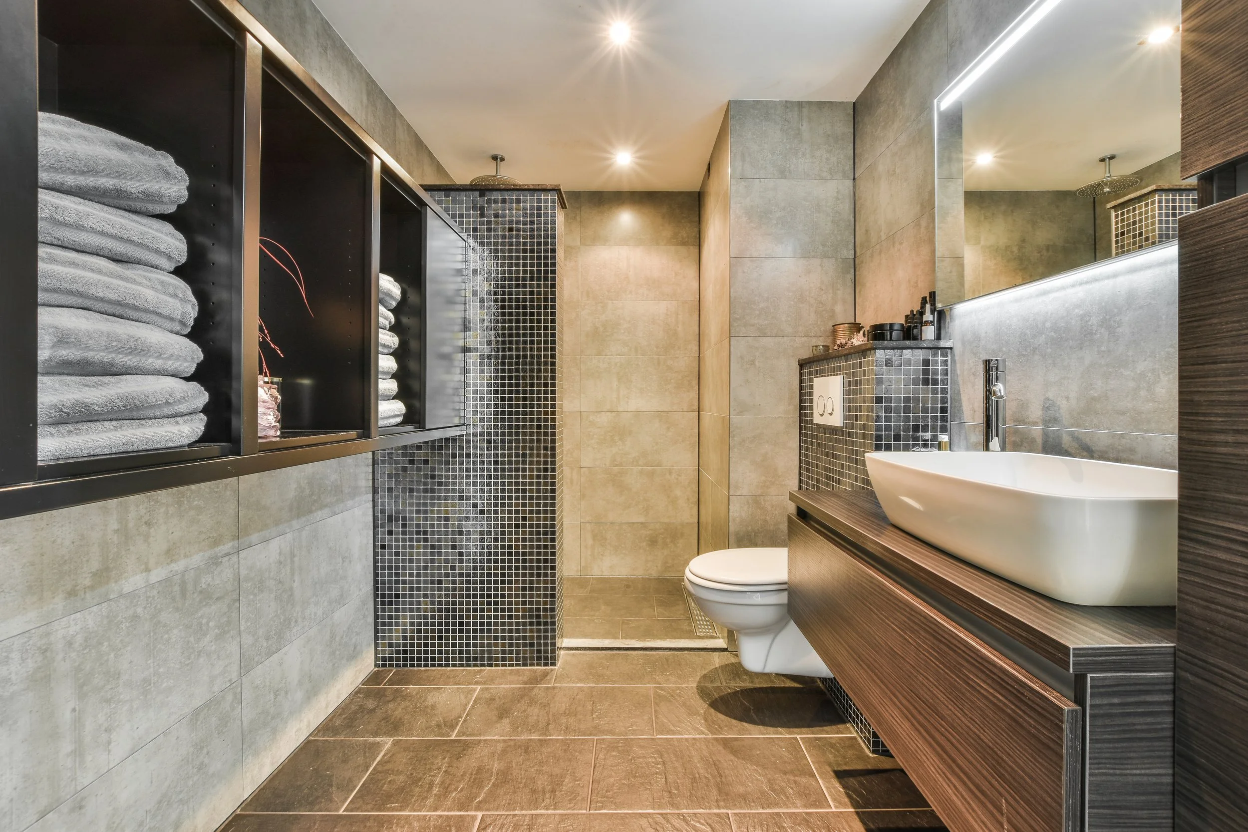 Modern bathroom with beige and brown tiles, a floating wooden vanity with a white vessel sink, wall-mounted faucet, toilet, black shelving with folded towels, glass shower with black tile accents, ceiling-mounted showerheads, and recessed lighting.