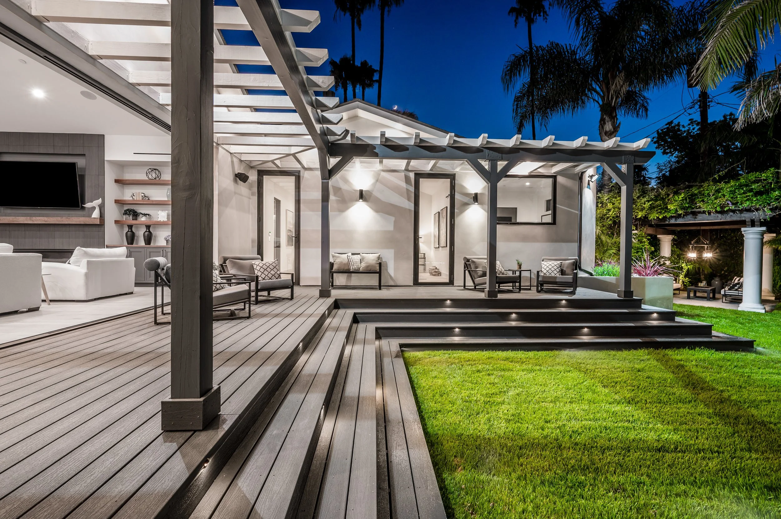Exterior view of a modern backyard with a wooden patio, outdoor furniture, and a pergola, surrounded by lush greenery and tall palm trees, during night time.