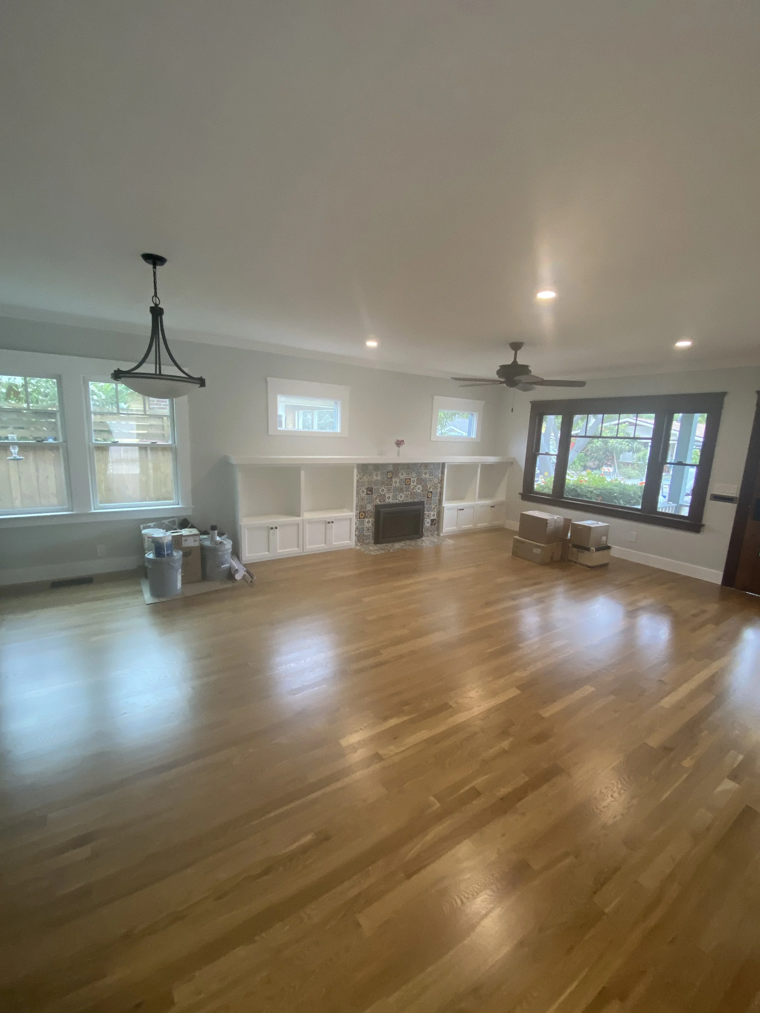 A living room with hardwood floors, white cabinets near a fireplace, multiple windows, and both a ceiling fan and a chandelier. Some boxes are on the floor.