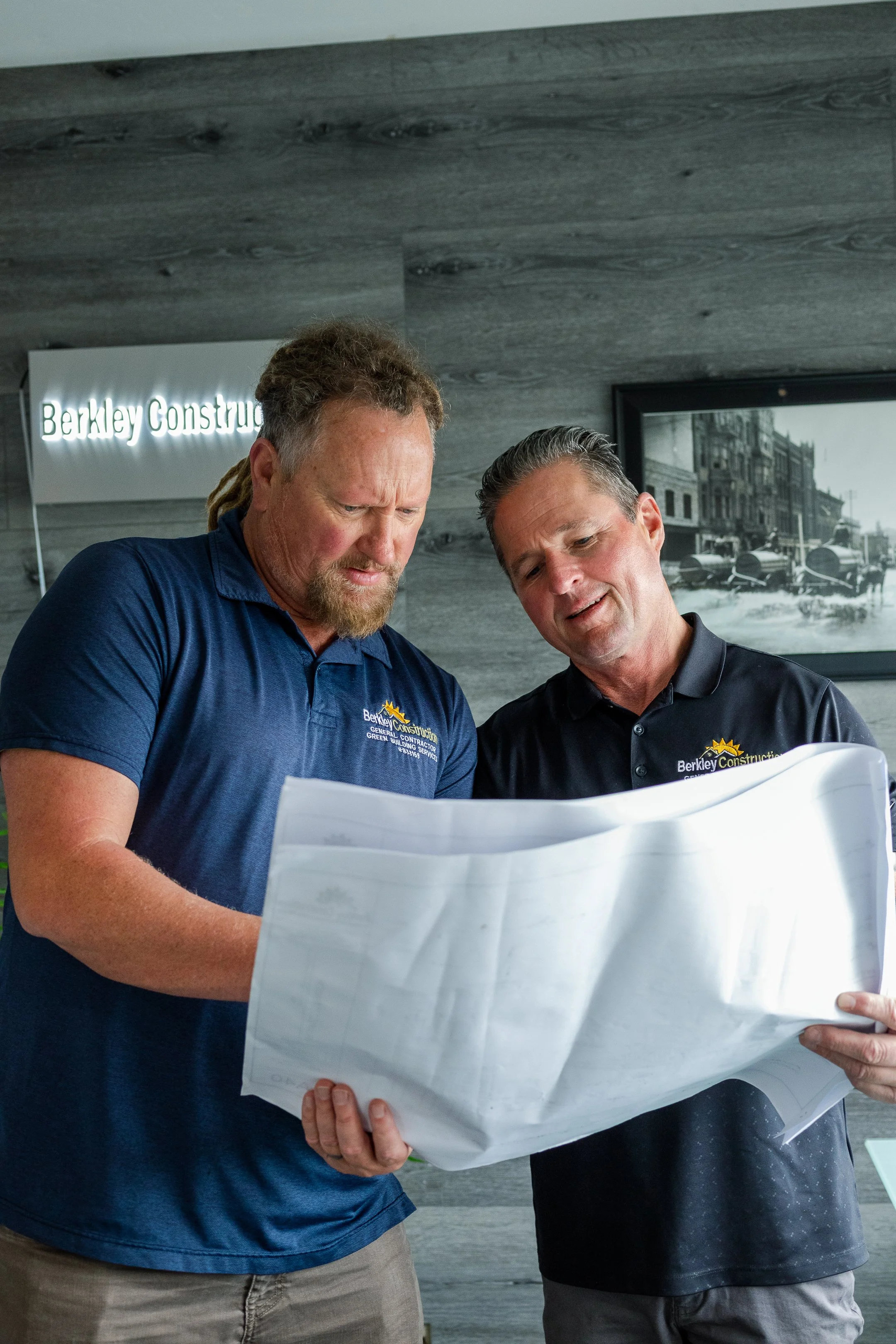 Two men reviewing detailed blueprints or construction plans inside an office or conference room, with a sign that reads 'Berkley Construction' and a historical black and white picture on the wall.