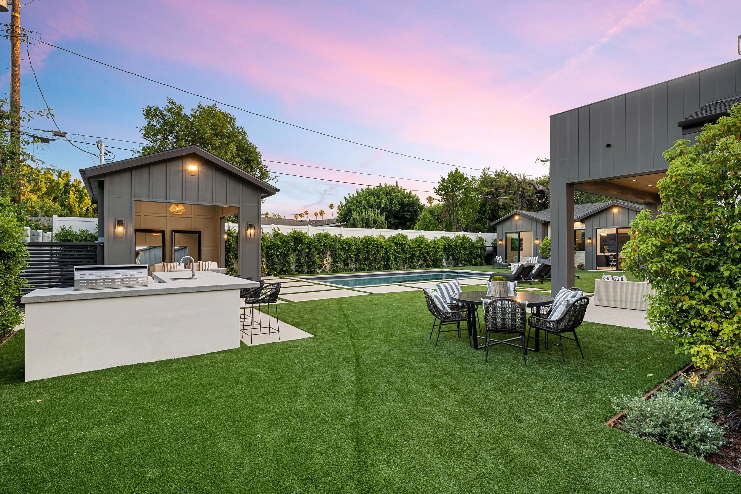 A modern backyard with a swimming pool, outdoor seating, and a small covered lounge area. The yard is landscaped with a green lawn, trees, and shrubbery under a colorful sunset sky.