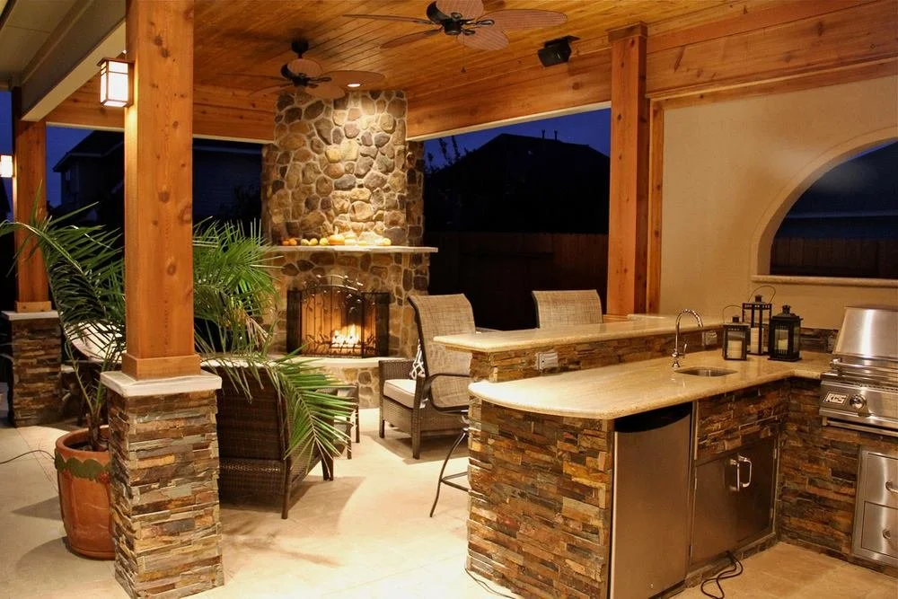 Outdoor covered patio area with a stone fireplace, seating area with woven chairs and a couch, and a kitchen counter with a sink, lanterns, and a grill, illuminated by warm lighting