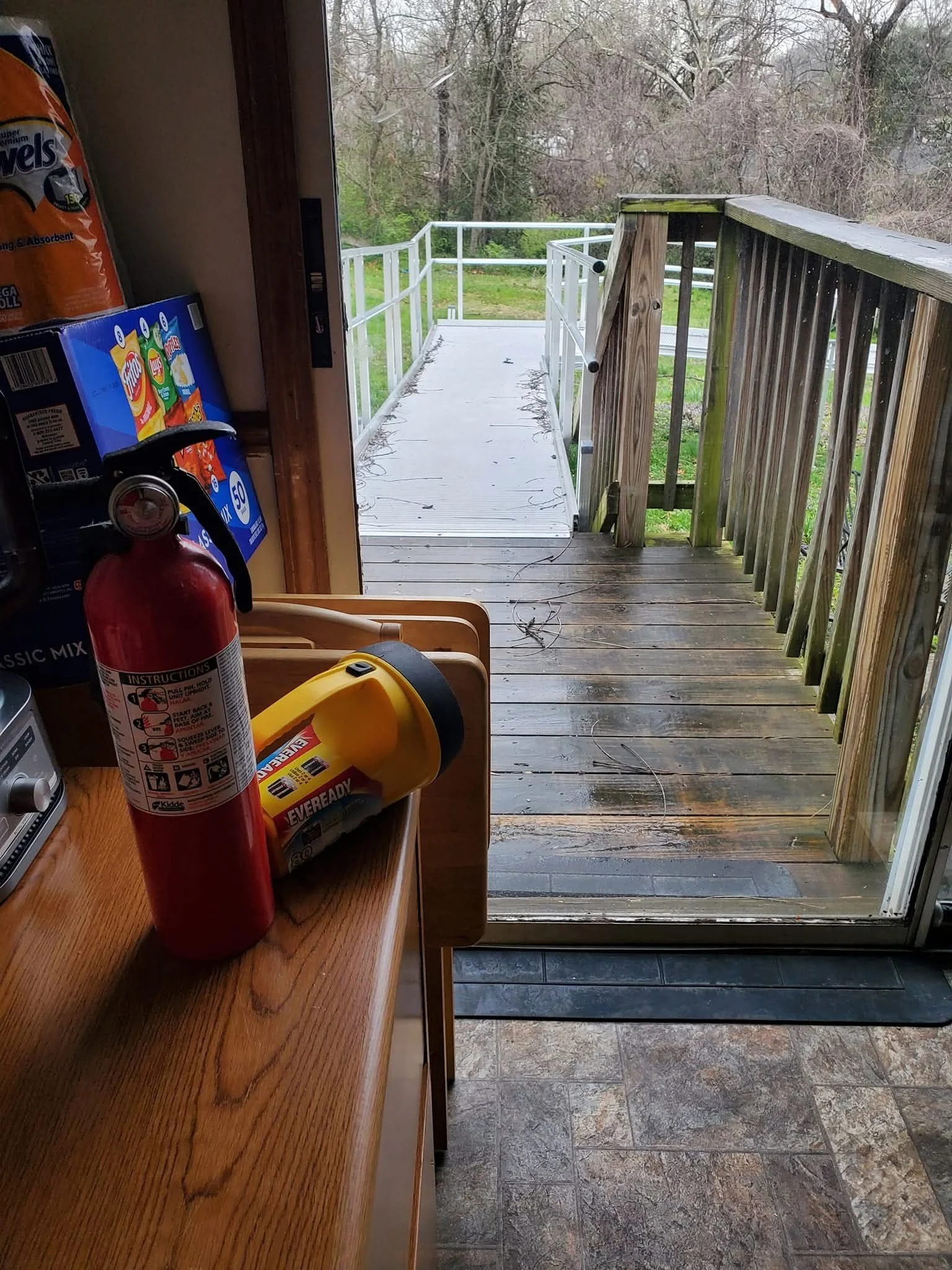 View of a wooden deck outside a sliding glass door from inside a kitchen. Inside, there is a red fire extinguisher, a yellow container of garbage bags, and some kitchen items on a wooden counter. The deck has a white metal railing and looks out onto 