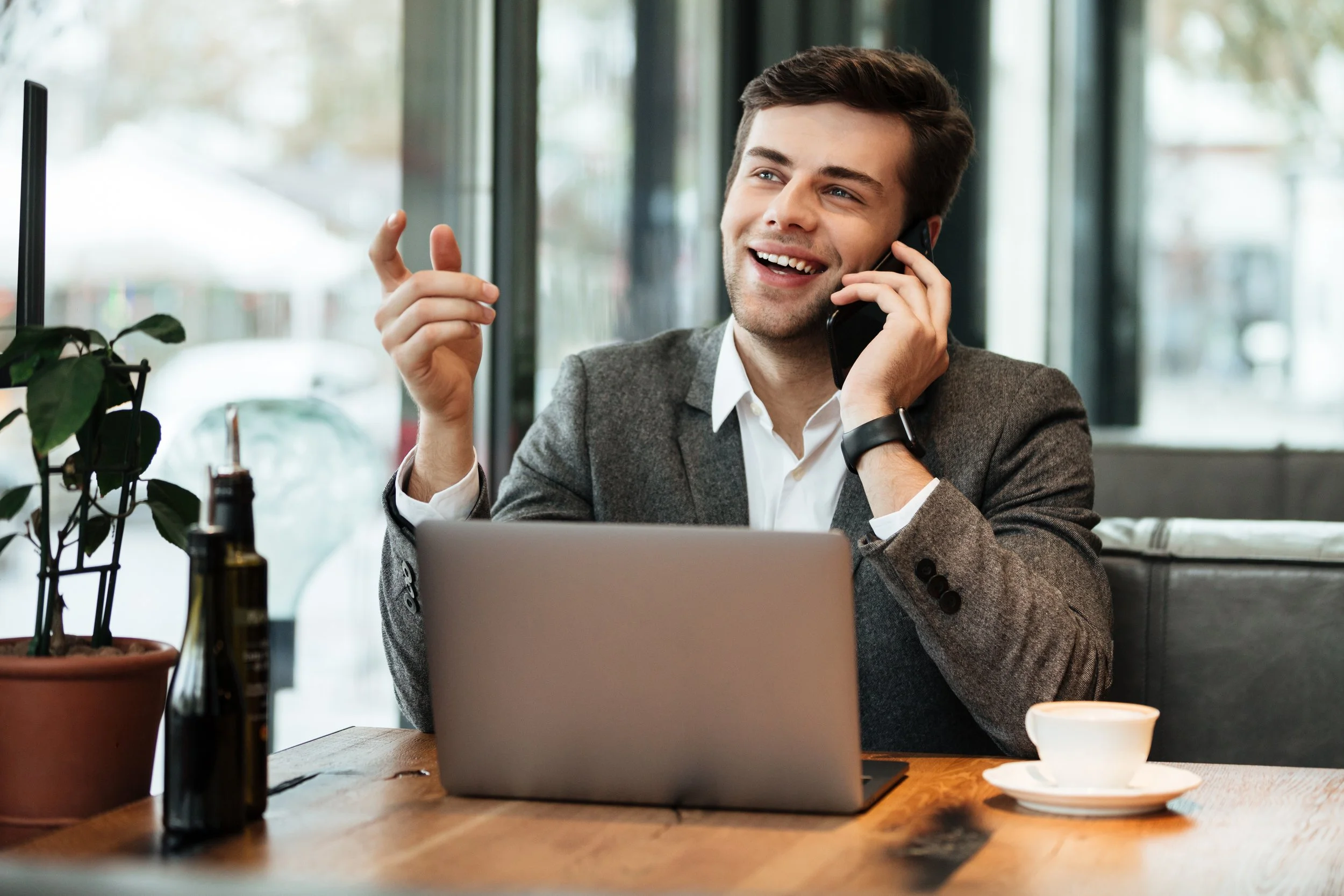 Man in a gray blazer talking on a cell phone, smiling, sitting at a wooden table with a laptop, a coffee cup, a bottle, and a potted plant in a cafe.