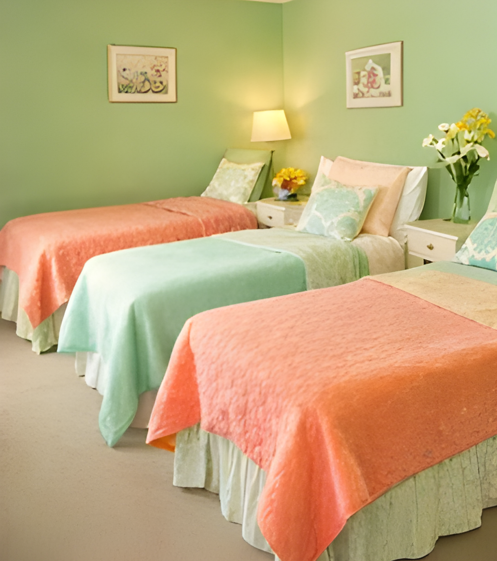 Three single beds with pastel-colored bedding and pillows in a room with green walls, flower arrangements on side tables, and framed artwork on the walls.