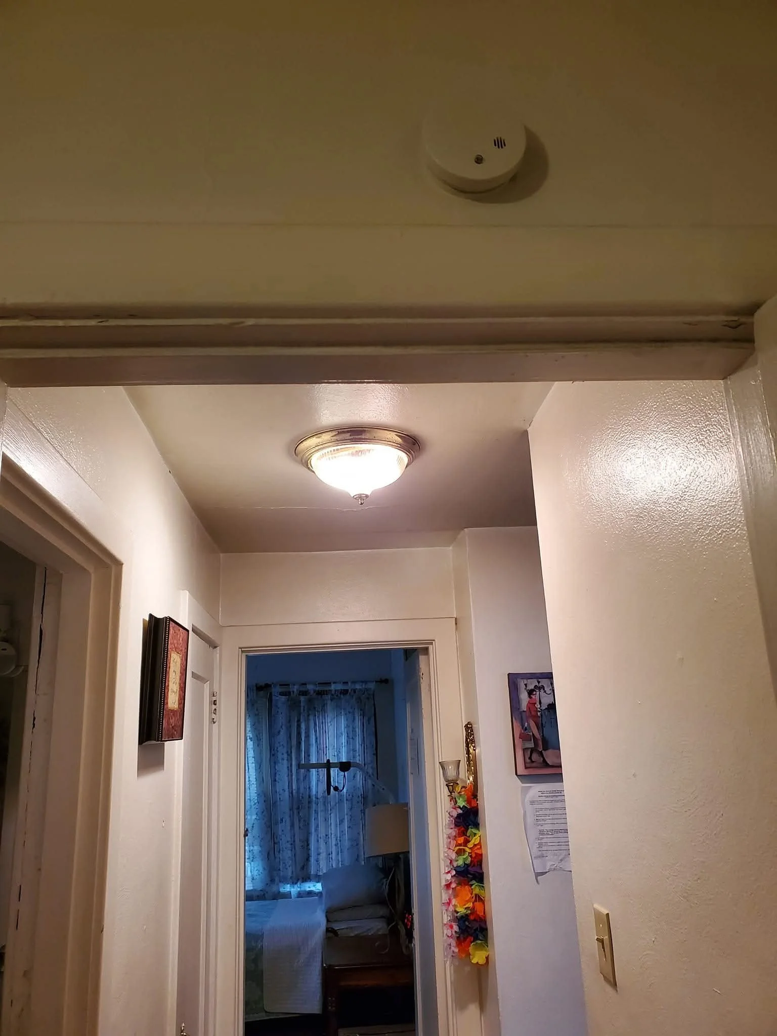 Hallway with ceiling light, doorway to a room with a bed and curtains, colorful hanging decoration, and framed artwork on the wall.