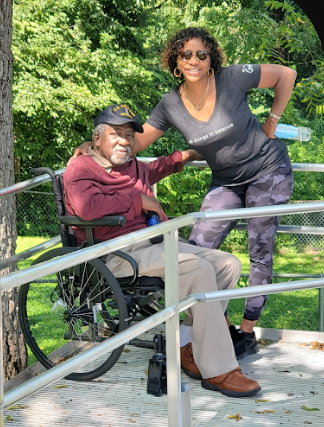 A woman with curly hair and sunglasses poses next to an elderly man in a wheelchair on a bridge during daytime, with green trees in the background.