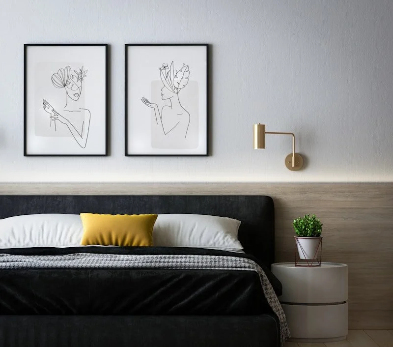 Modern bedroom with black bed frame, white bedding, a yellow pillow, wall art of line drawings of women, a wall lamp, and a small round white nightstand with a potted plant.
