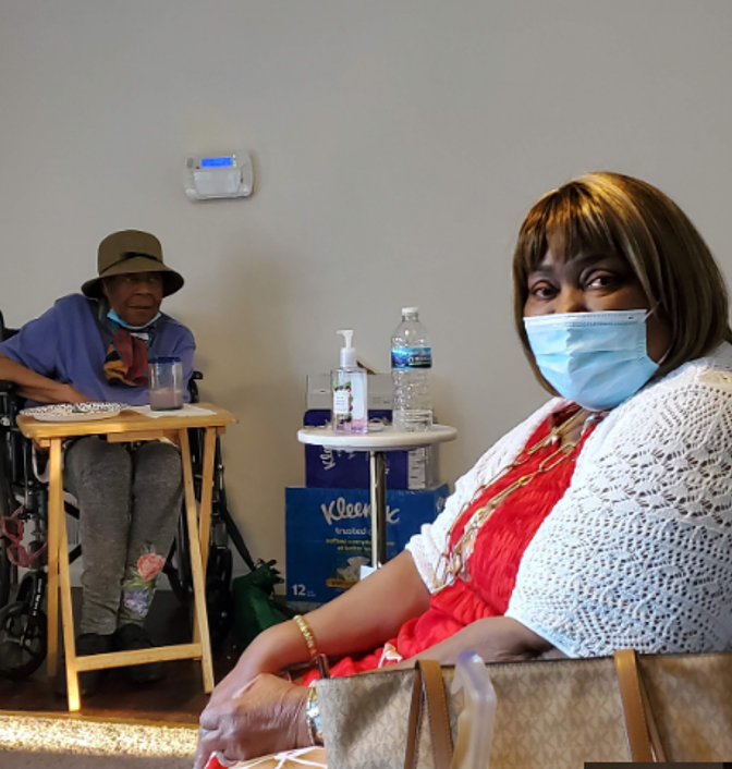 Two women, one wearing a face mask, are seated indoors. The woman in the foreground has short hair, is wearing a red top with a white shawl, and has a handbag on her lap. The woman in the background is sitting at a table, wearing a hat, glasses, and 