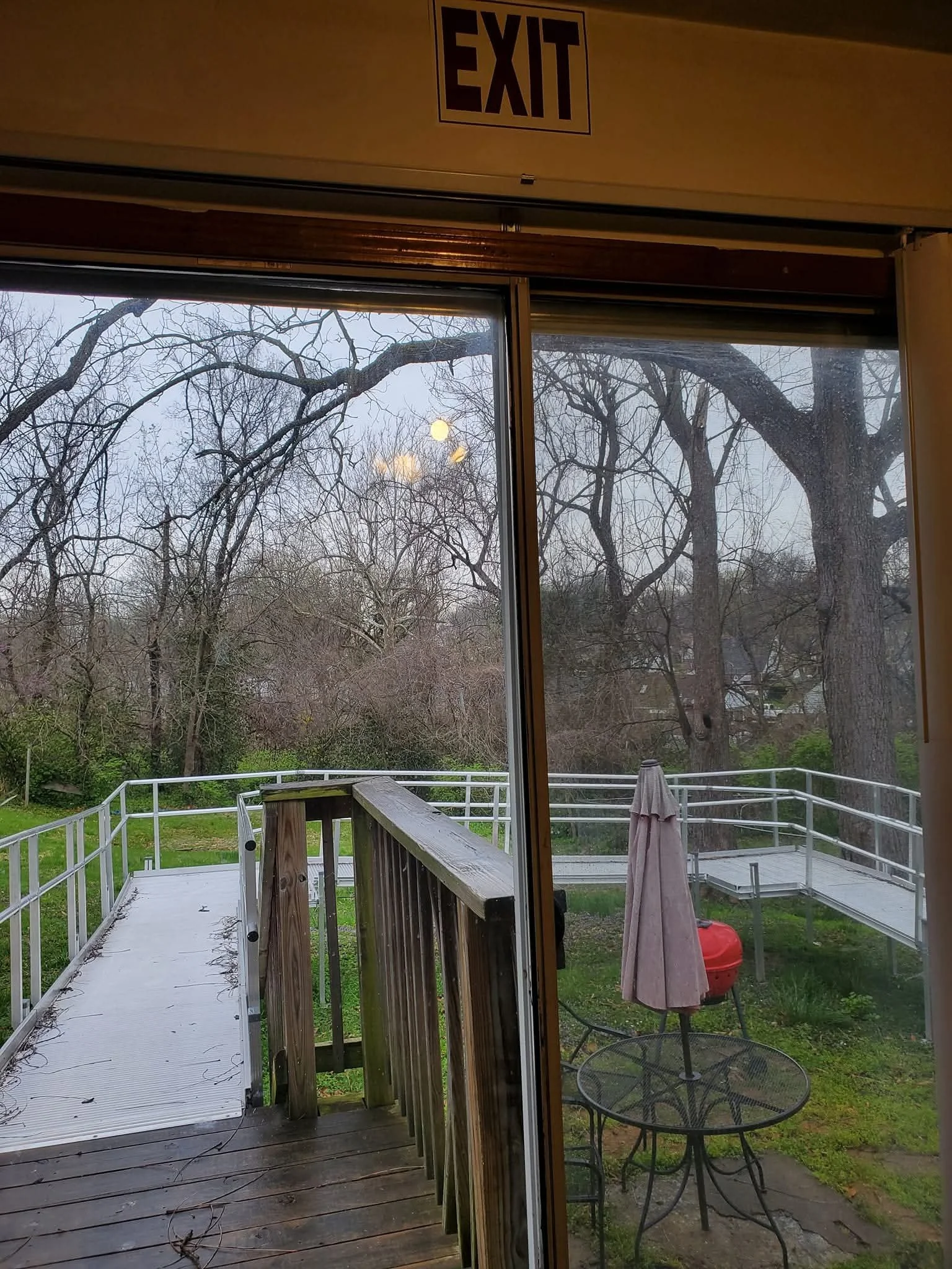 View through a sliding glass door showing a wooden deck with a metal railing, a patio table with an umbrella, a red grill, several leafless trees, and a cloudy sky.