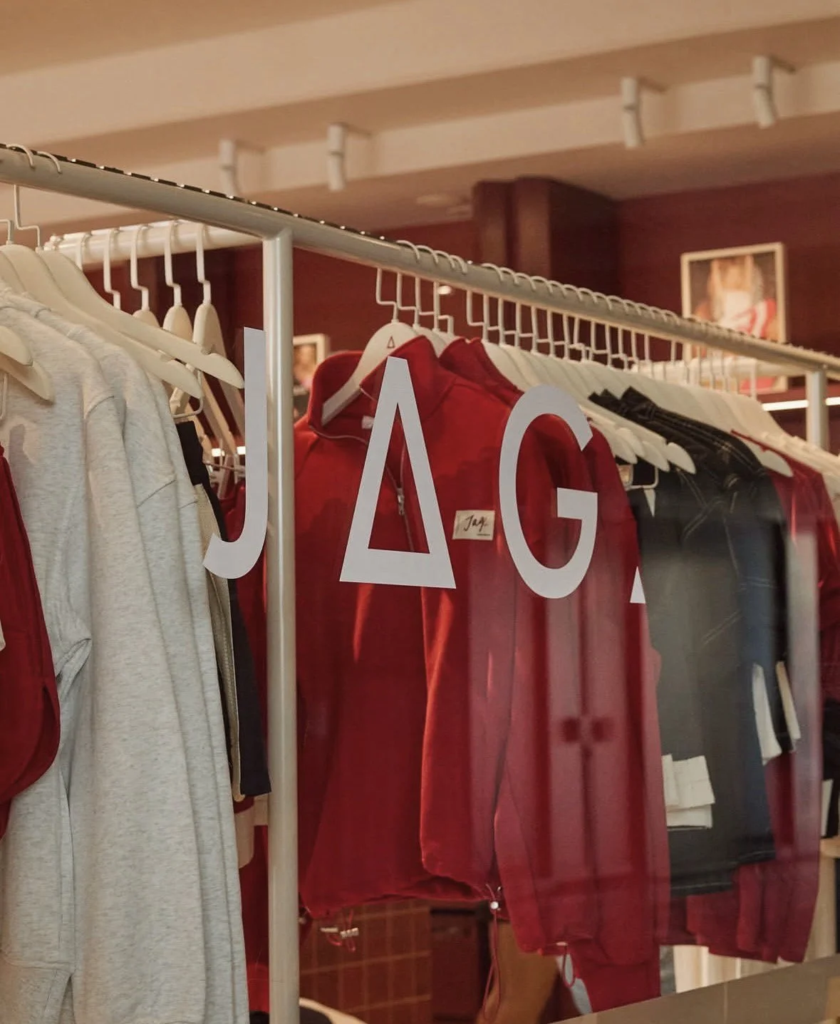 Clothing rack displaying various garments, including a red jacket and a gray hoodie, with white Hang tags, in a store or boutique. JAG Bondi Beach Flagship store.