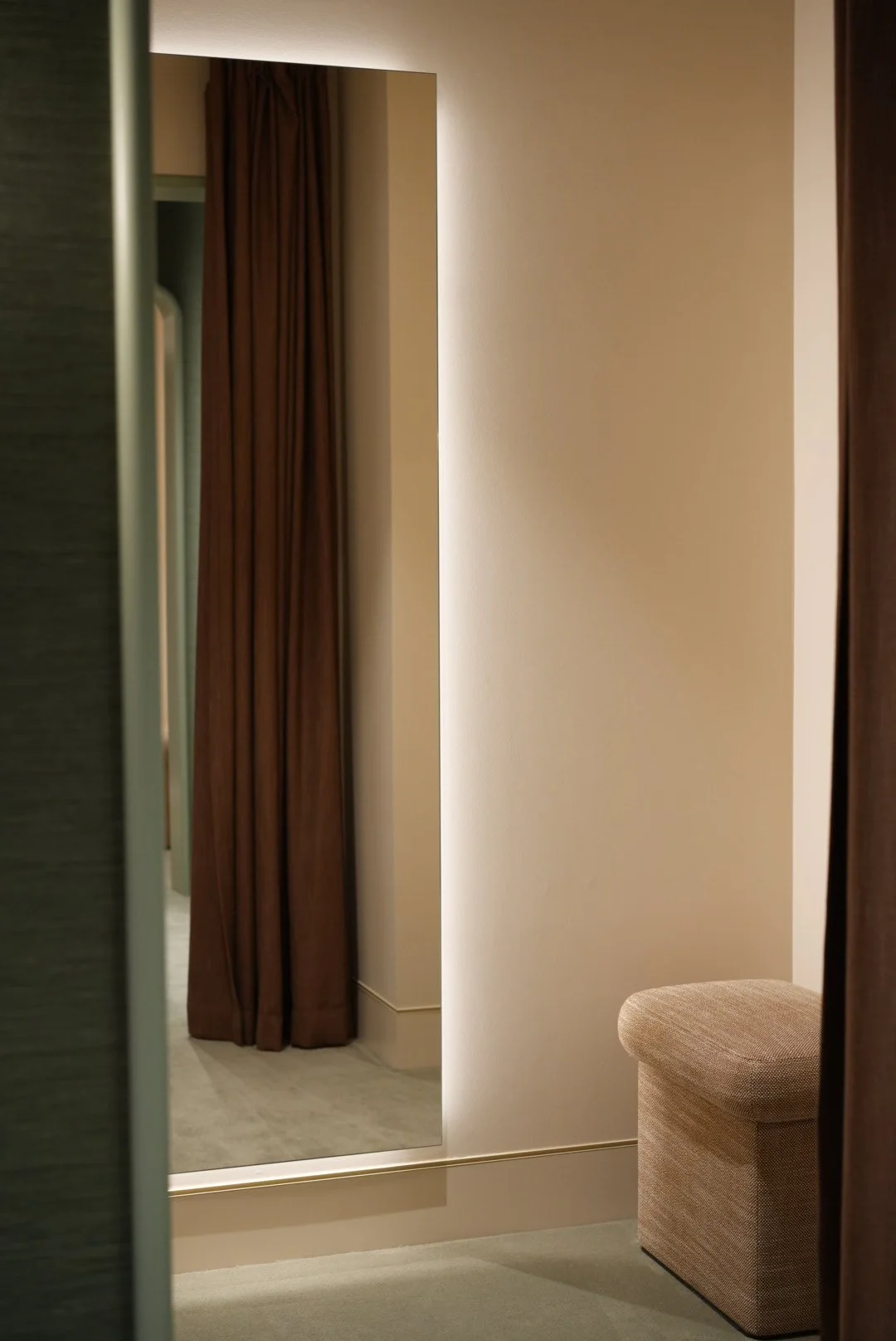 Dressing room interior with a large mirror, a brown curtain, beige wall, and beige upholstered ottoman.