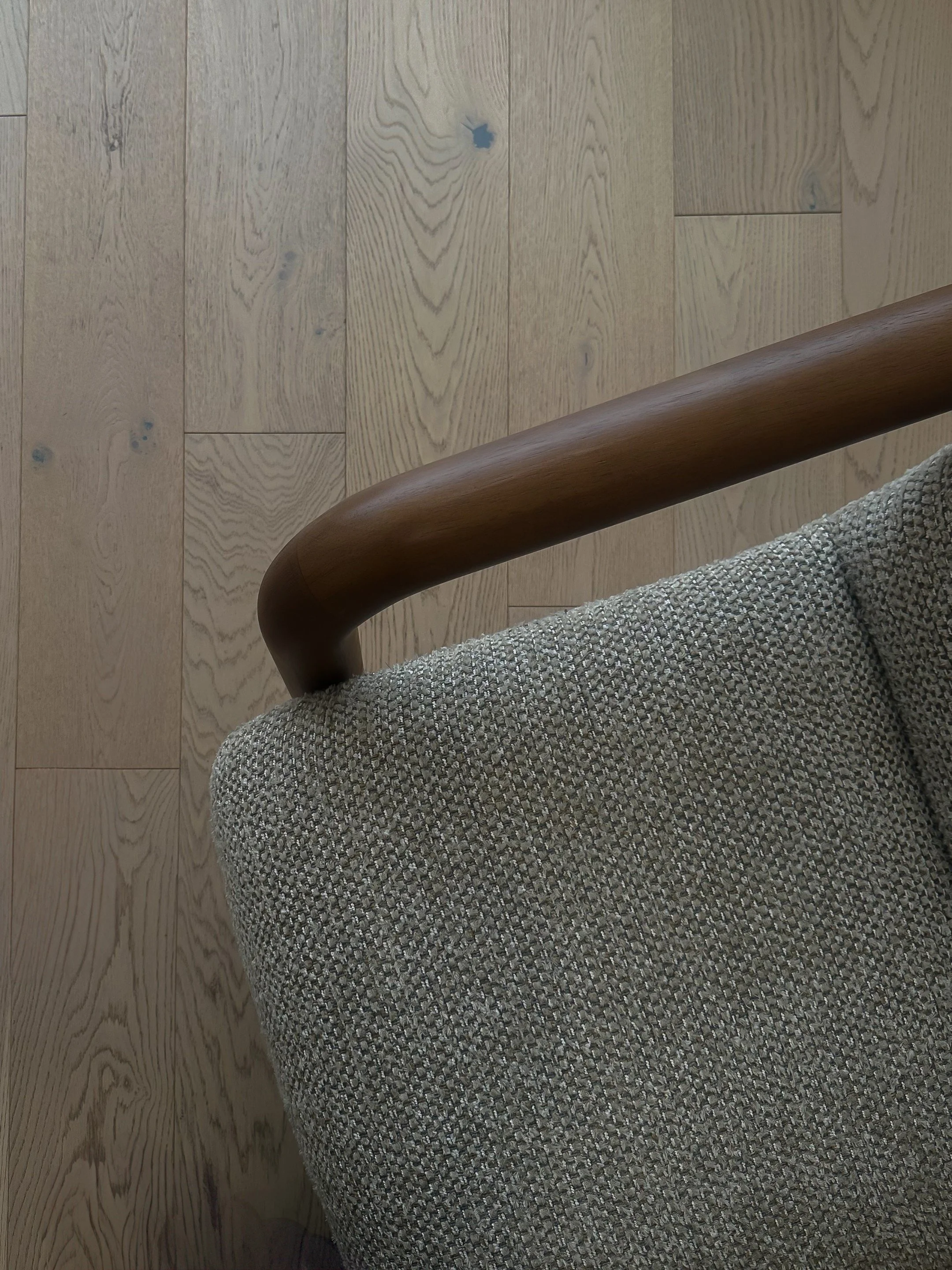 Close-up of a wooden armrest attached to a fabric-covered chair on a wooden floor.