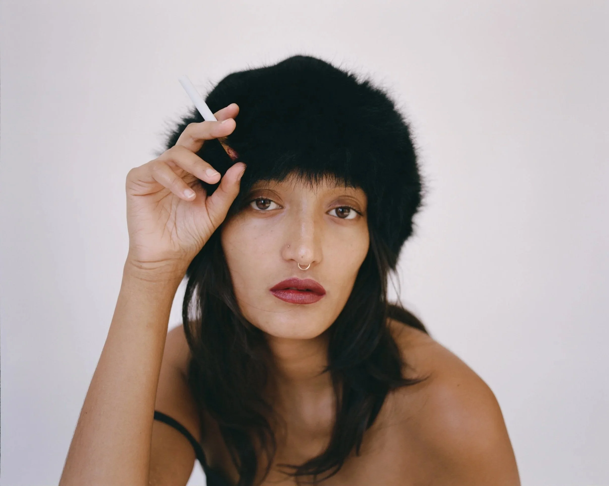 Woman with dark hair wearing a black furry hat, holding a cigarette near her temple, against a plain white background.