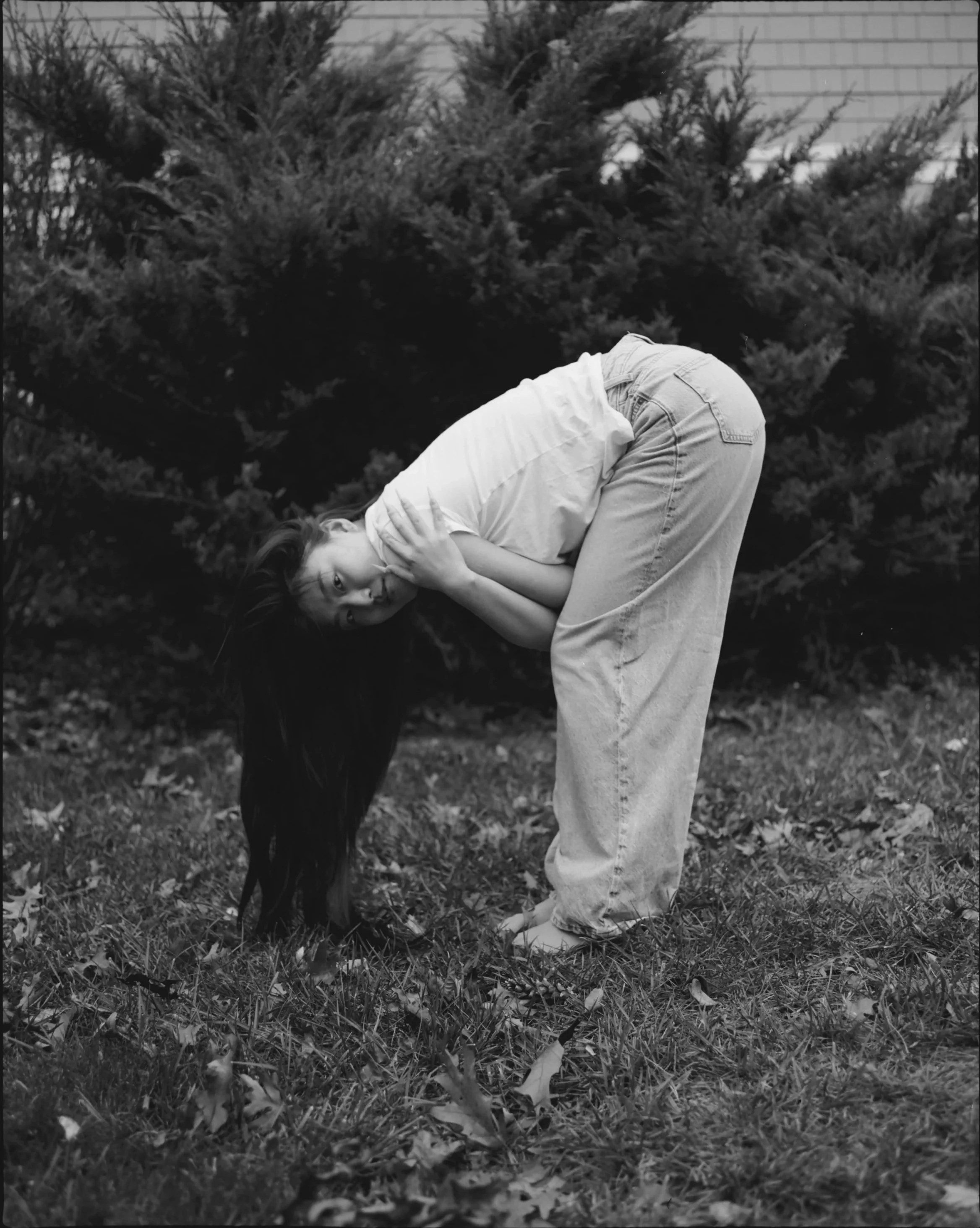 A woman bending over with her hair hanging down, holding her neck with both hands, standing on grass with bushes in the background.