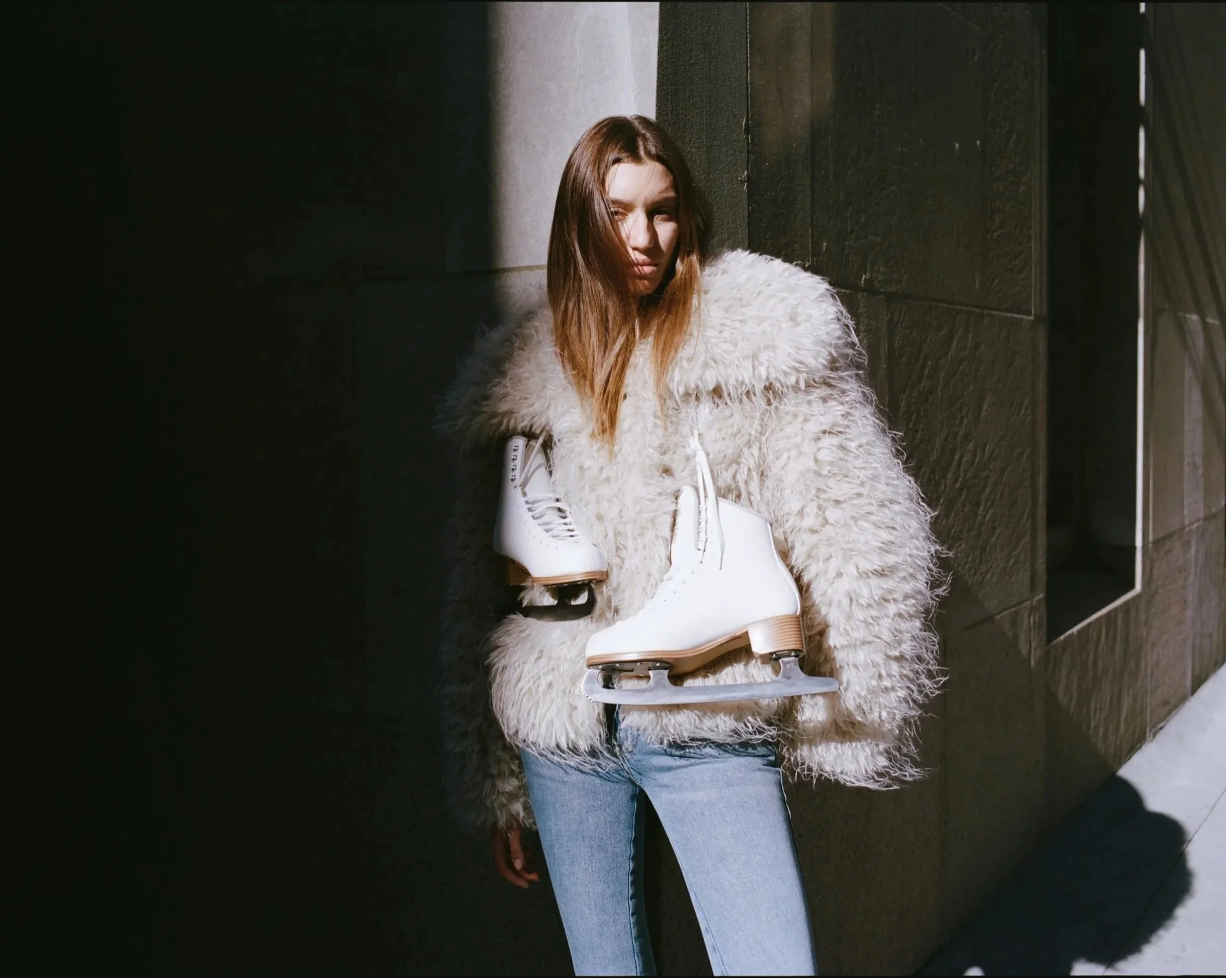 A woman with long brown hair wearing a fluffy white coat and blue jeans, holding the ice skates in front of her, standing near dark gray stone walls in bright sunlight.