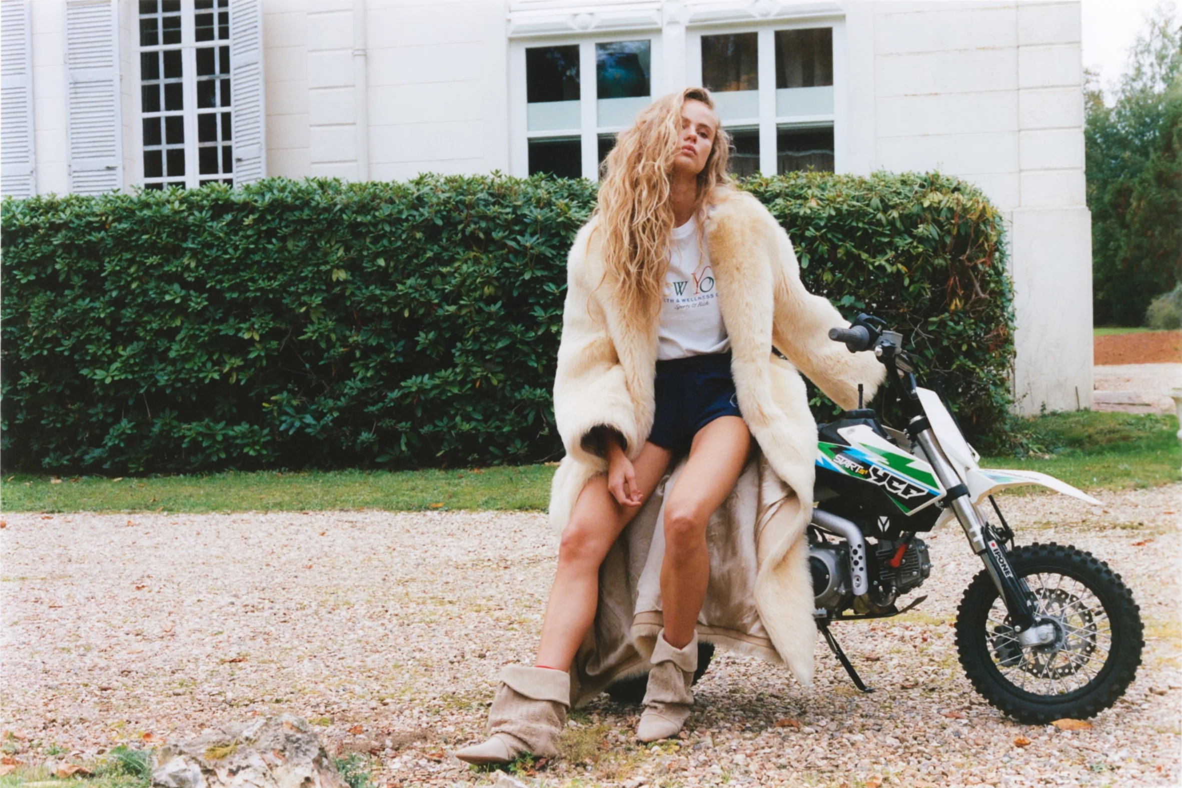 Young woman with long blonde hair, wearing a white t-shirt, dark shorts, and a beige faux fur coat, sitting on a small dirt bike outdoors in front of a white house with green bushes.