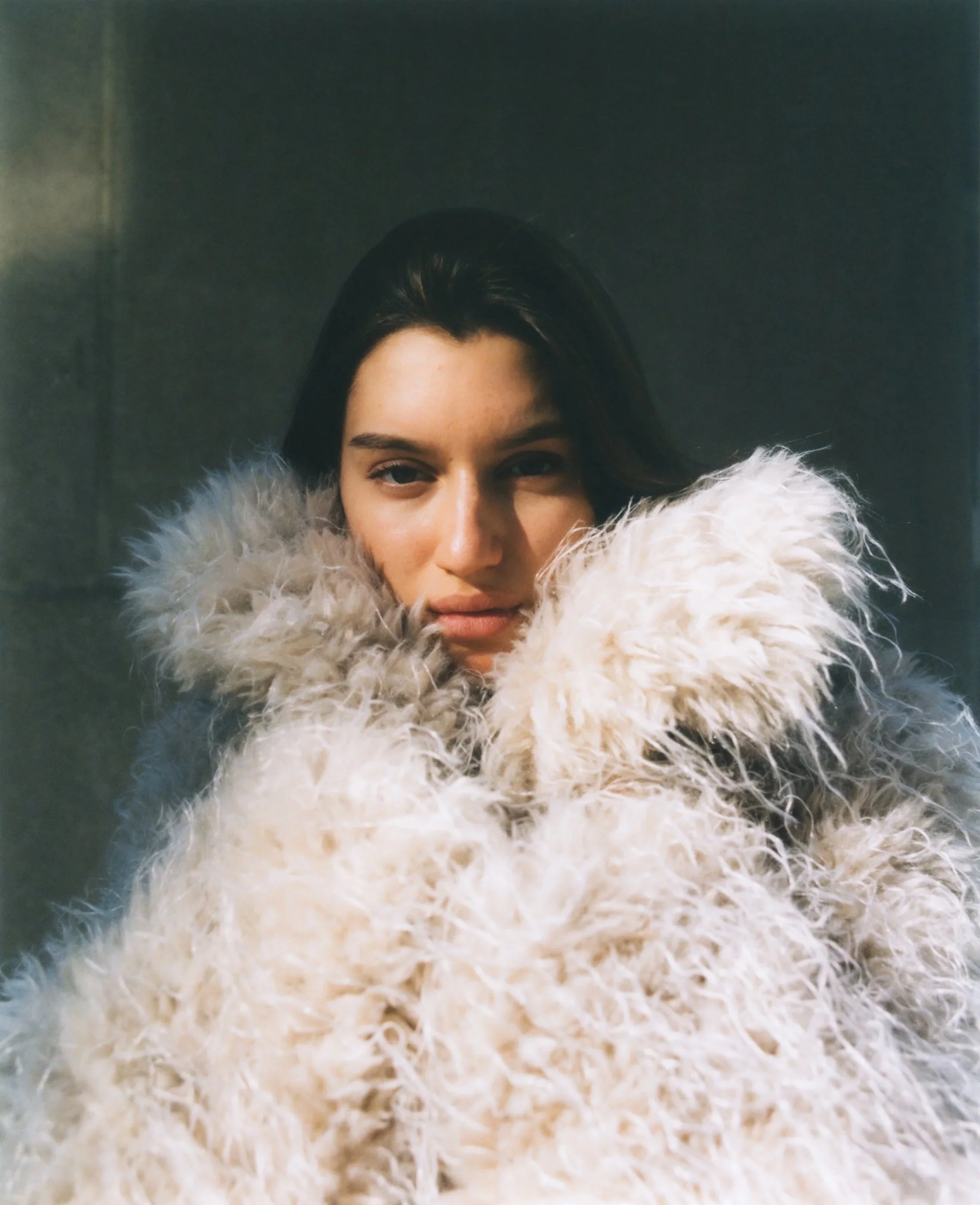 A woman with dark hair wearing a large, fluffy, cream-colored faux fur coat or stole, standing against a gray wall.