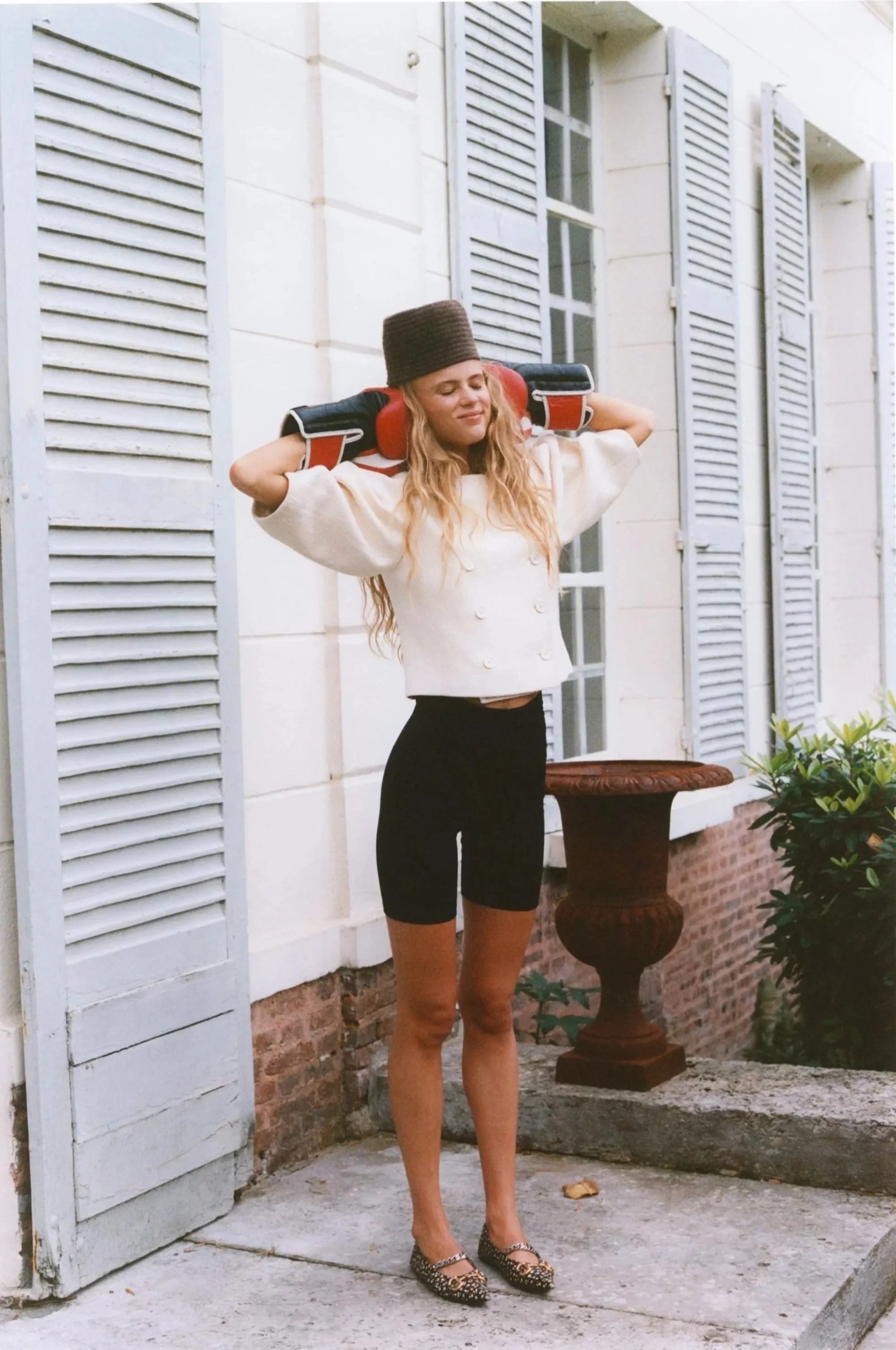 A woman standing outside wearing black bike shorts, a white cropped double-breasted jacket, leopard print flats, with her eyes closed and hands resting on a helmet on her shoulders.
