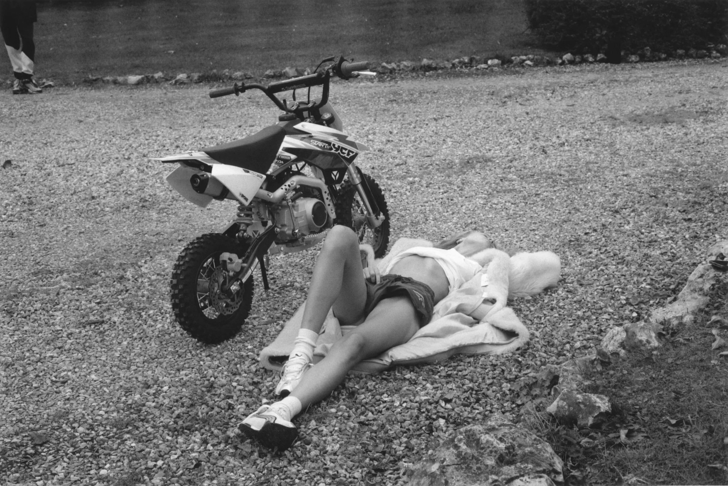 A person lying on a towel on a gravel surface next to a dirt bike, in an outdoor setting.