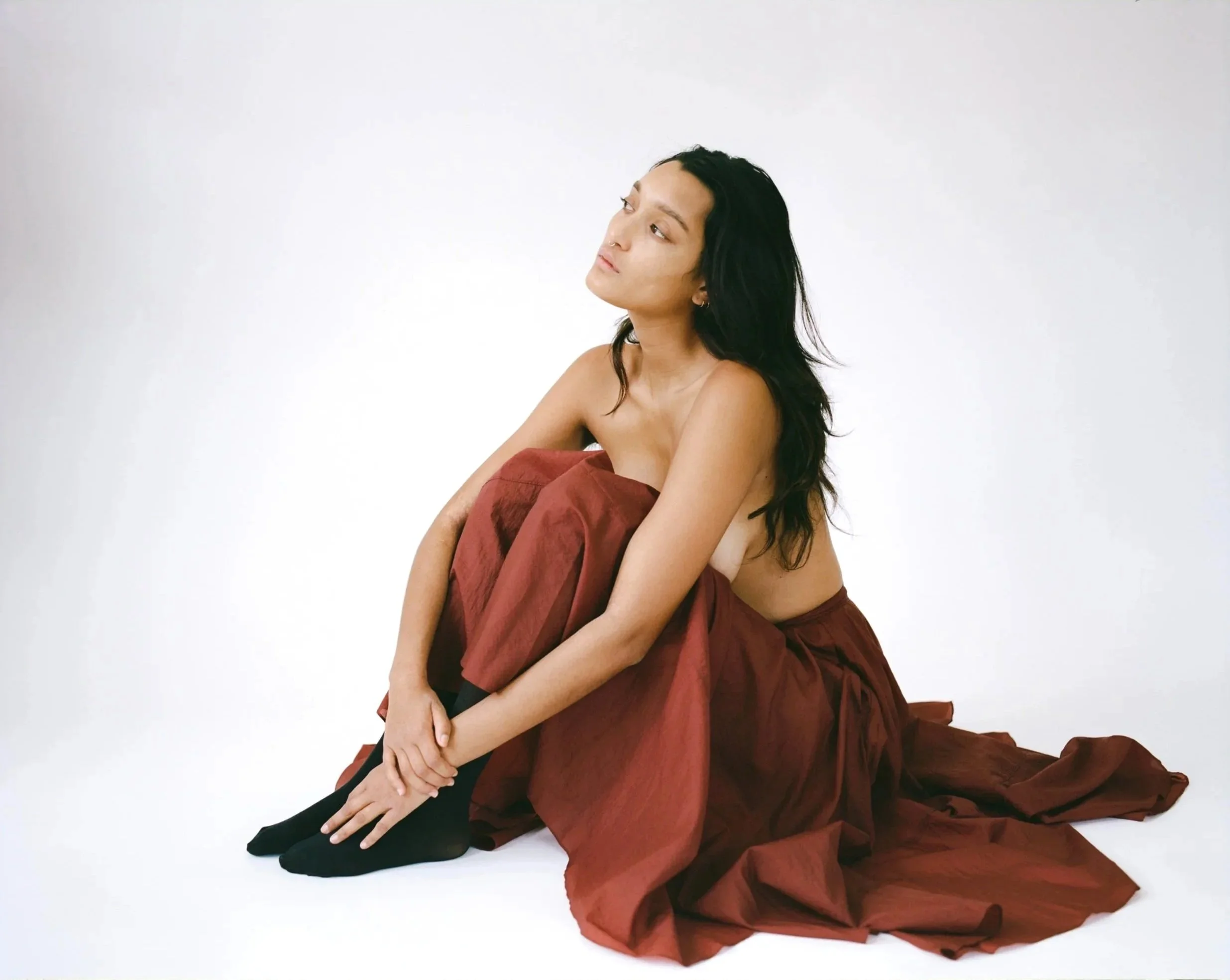 A woman with long black hair, sitting on the floor, wearing a long, dark red skirt, black shoes, and black socks, against a plain white background.