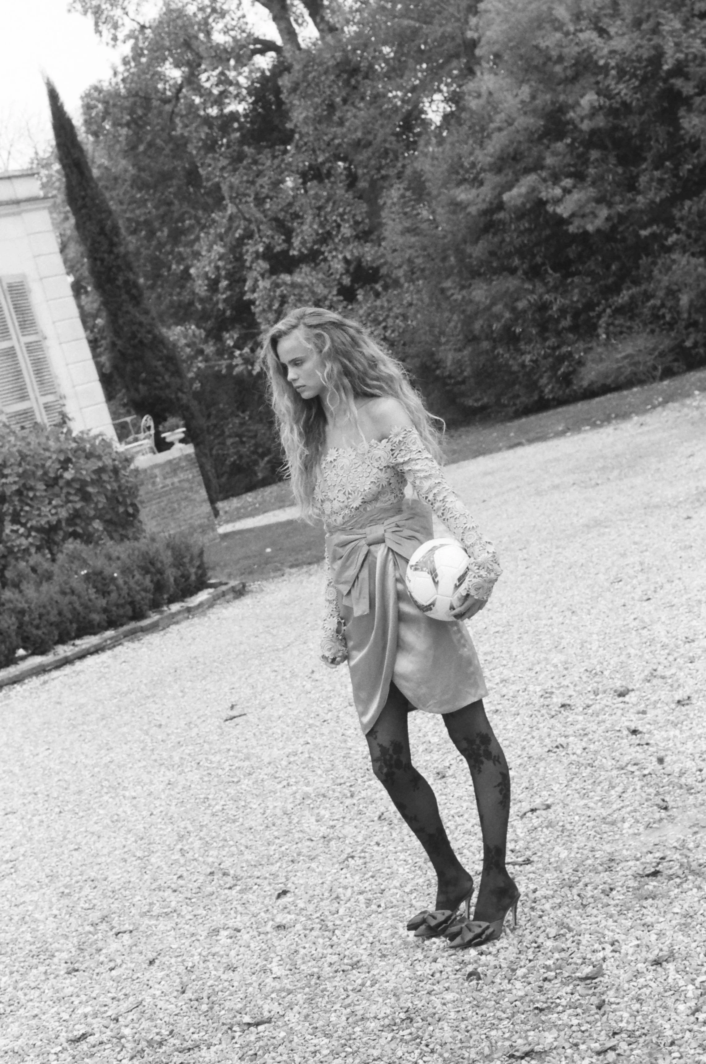 A woman standing outdoors on a gravel path, holding a soccer ball, wearing a lace top, skirt with a bow, floral tights, and heels, with trees and a house in the background.