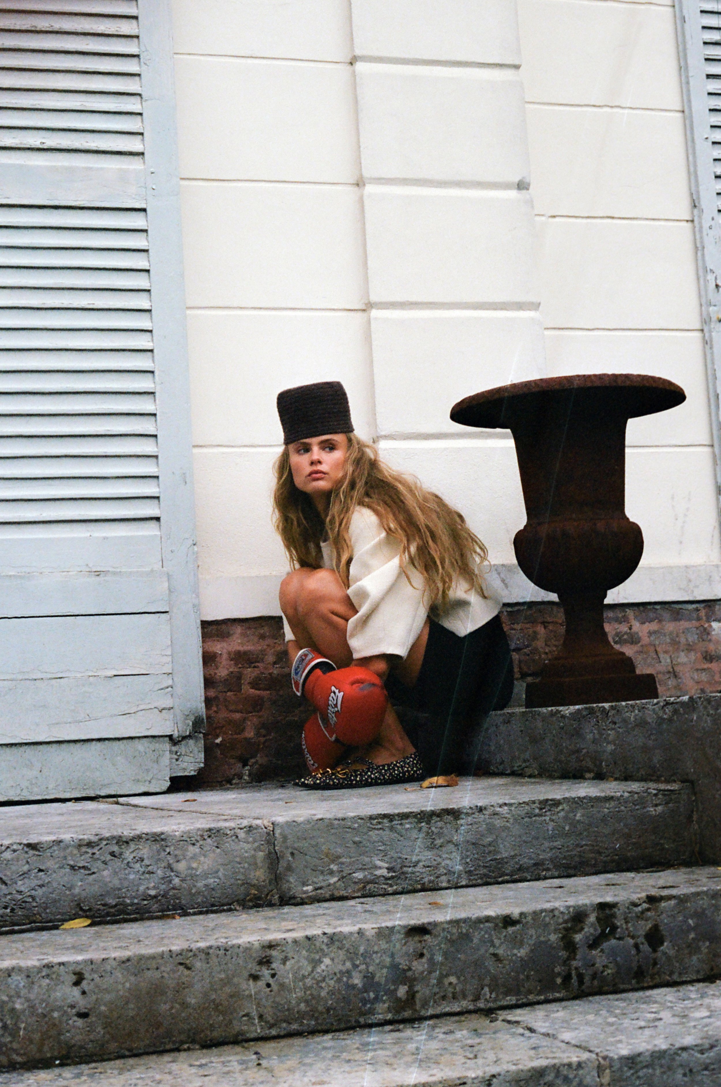 A young woman with long hair wearing a brown hat, white jacket, and red boxing gloves, crouching on steps outside a building with white walls and a decorative rusted urn.