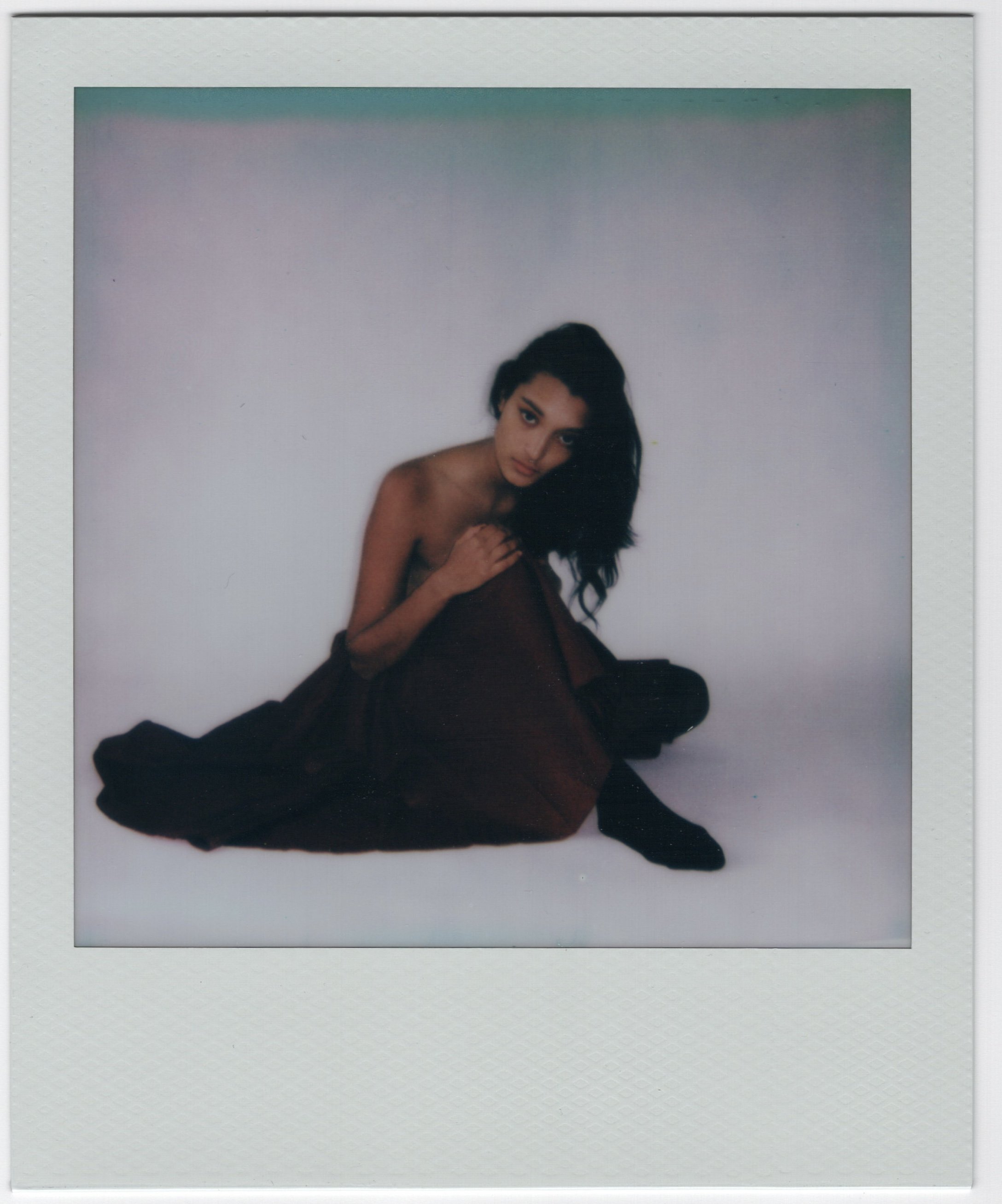 A woman sitting on the floor, draped in a dark brown or black fabric, with long dark hair and a neutral expression, against a plain white background.