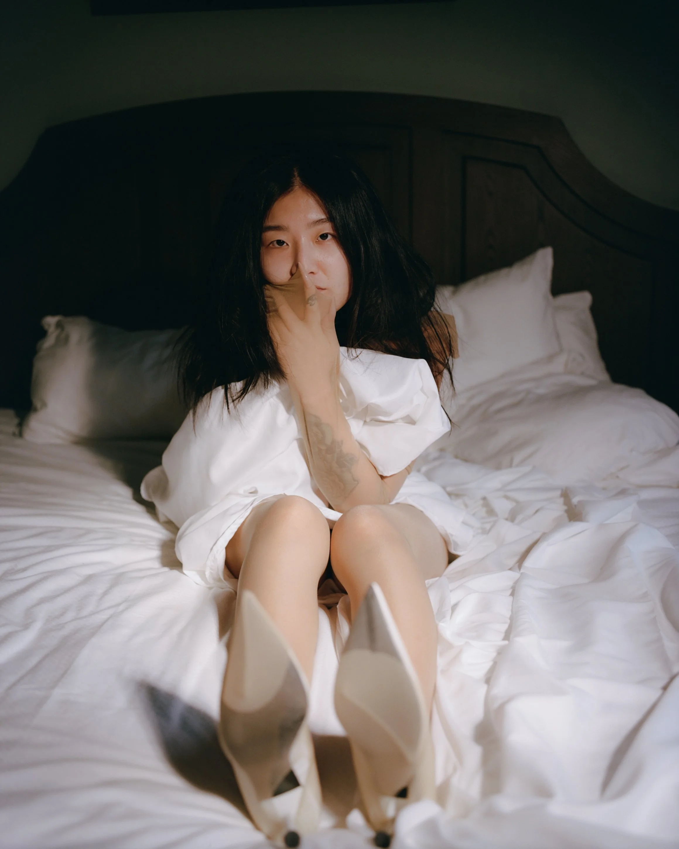A woman with black hair, sitting on a bed with white sheets, wearing white high heels and a white dress, with her legs extended toward the camera, making a playful gesture with her hand near her face.