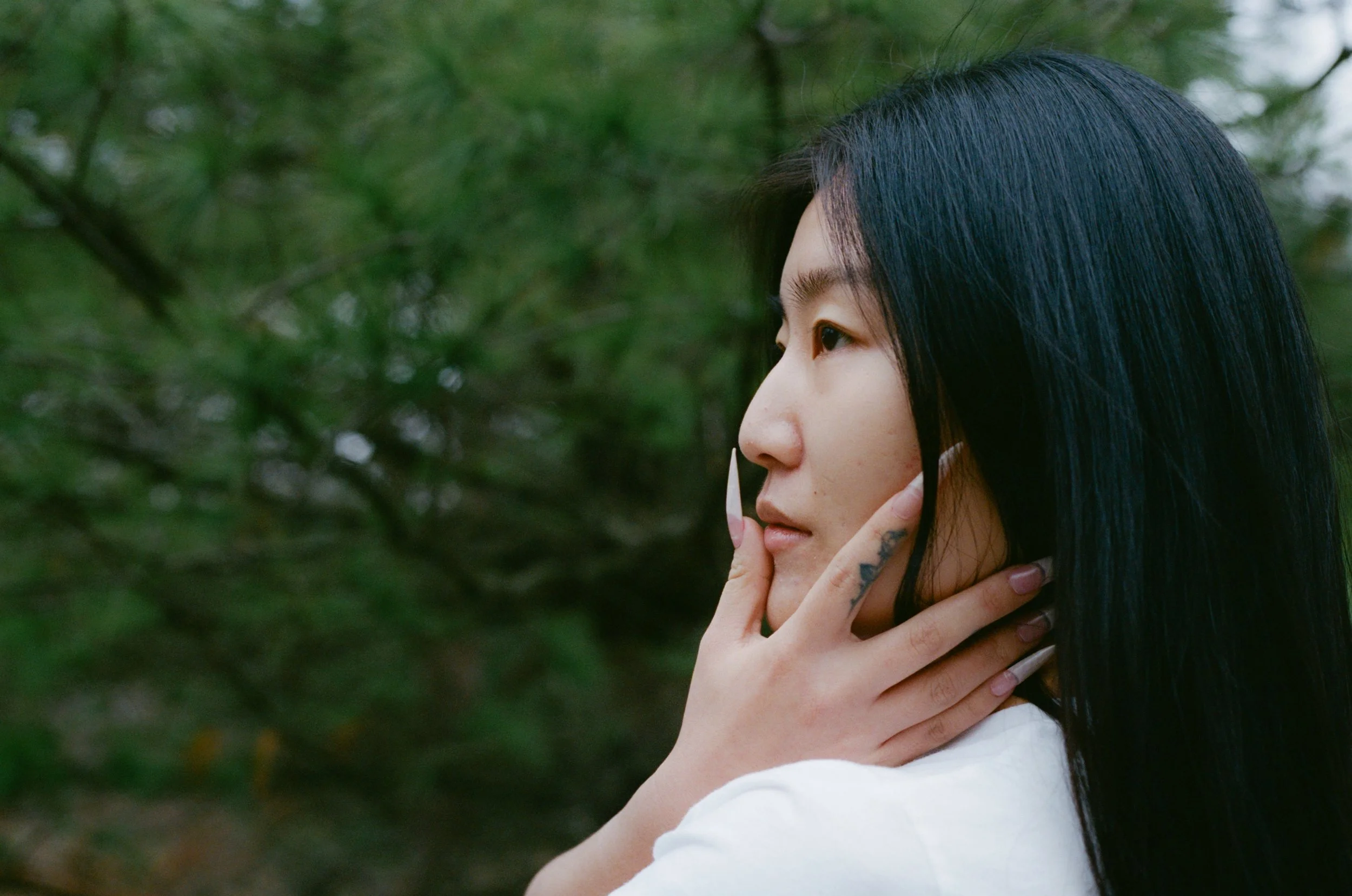 Profile of a young woman with long black hair, holding her chin with her hand, standing outdoors with green foliage in the background.