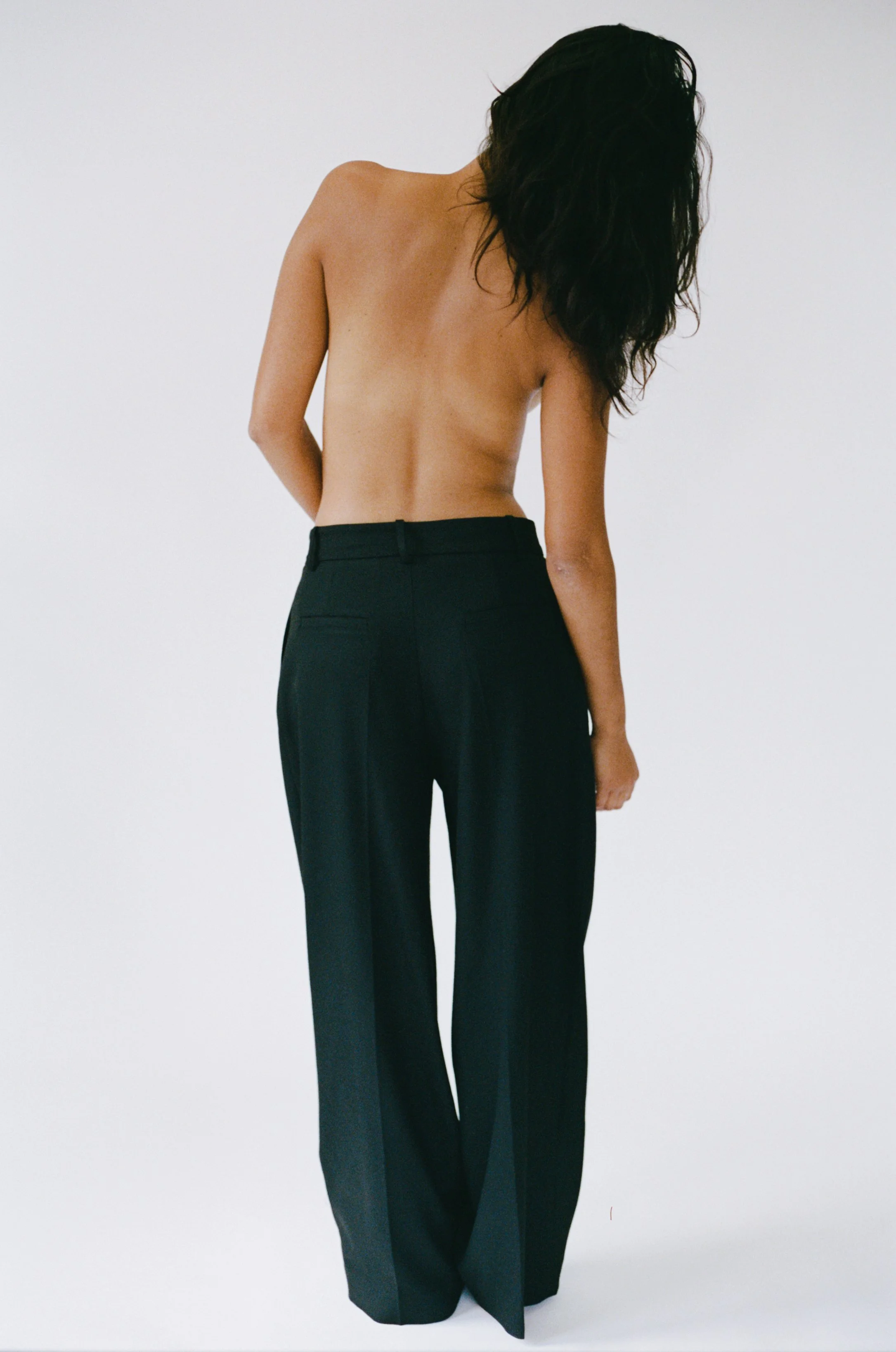 Back view of a topless woman with long dark hair wearing black pants, standing against a plain white background.