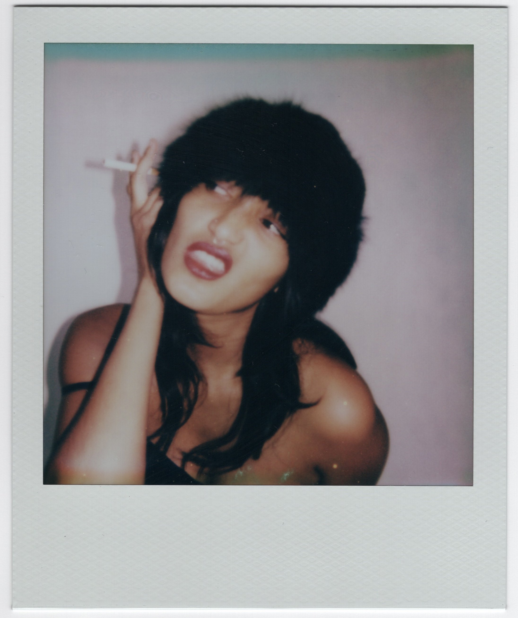 A young woman with dark, messy hair and red lipstick making a silly face, holding a cigarette in her left hand, against a light-colored wall.