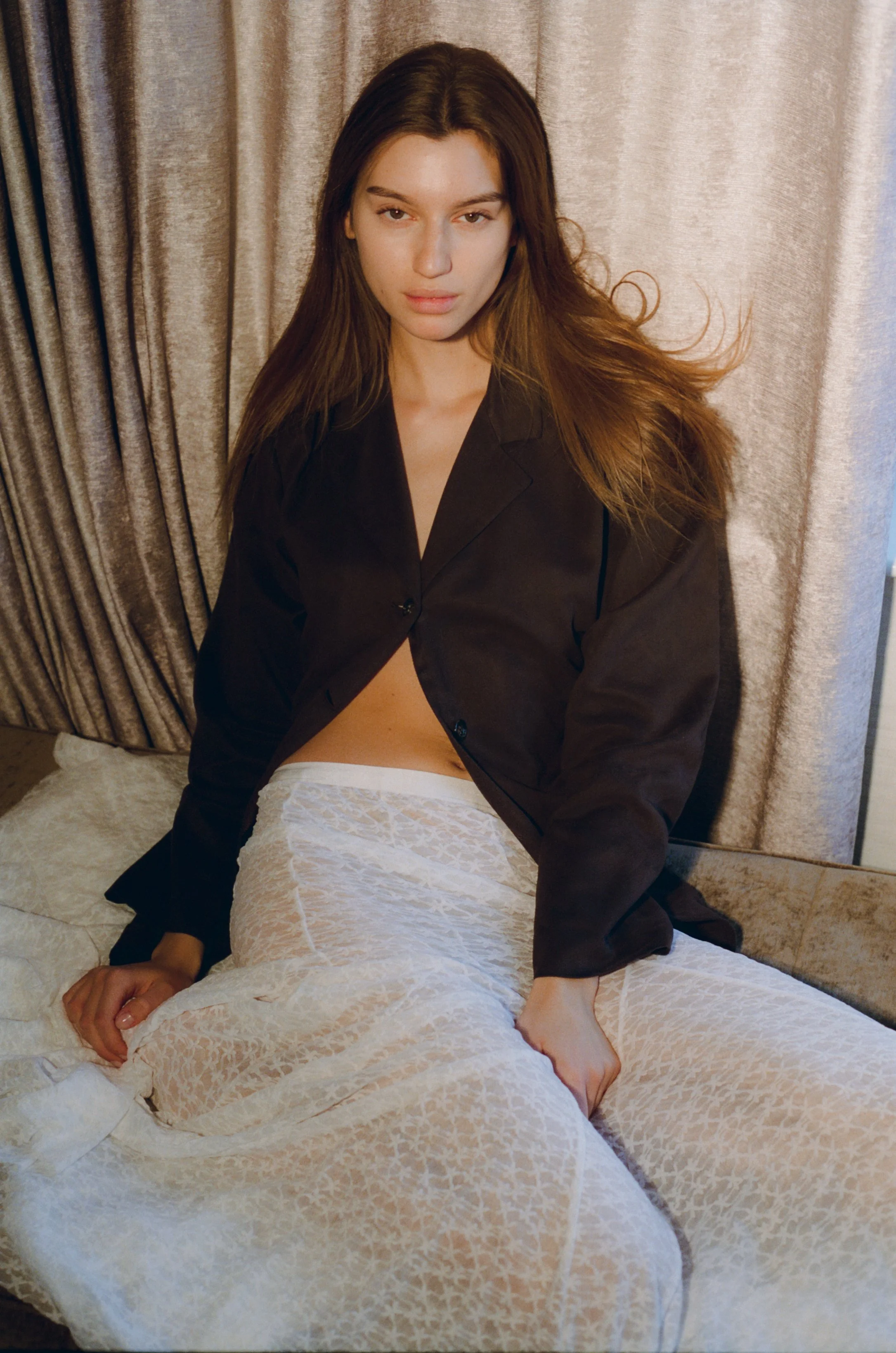 A young woman with long brown hair, wearing a black blazer and white lace dress, sitting on a bed with beige curtains in the background.