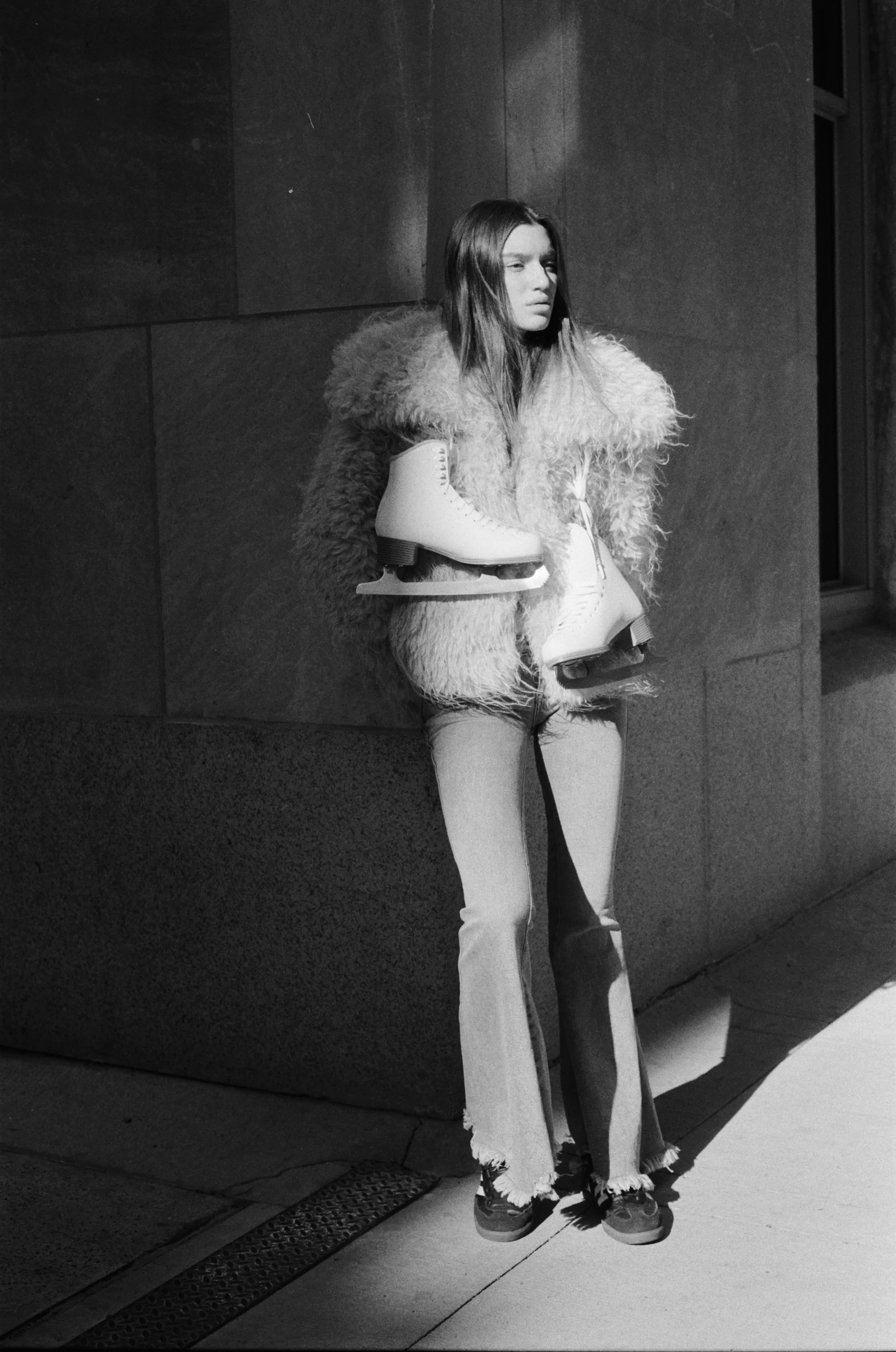 A young woman standing on a sidewalk at night, holding a pair of ice skates, wearing a fluffy coat, jeans, and sneakers, with a serious expression, illuminated by a streetlight.