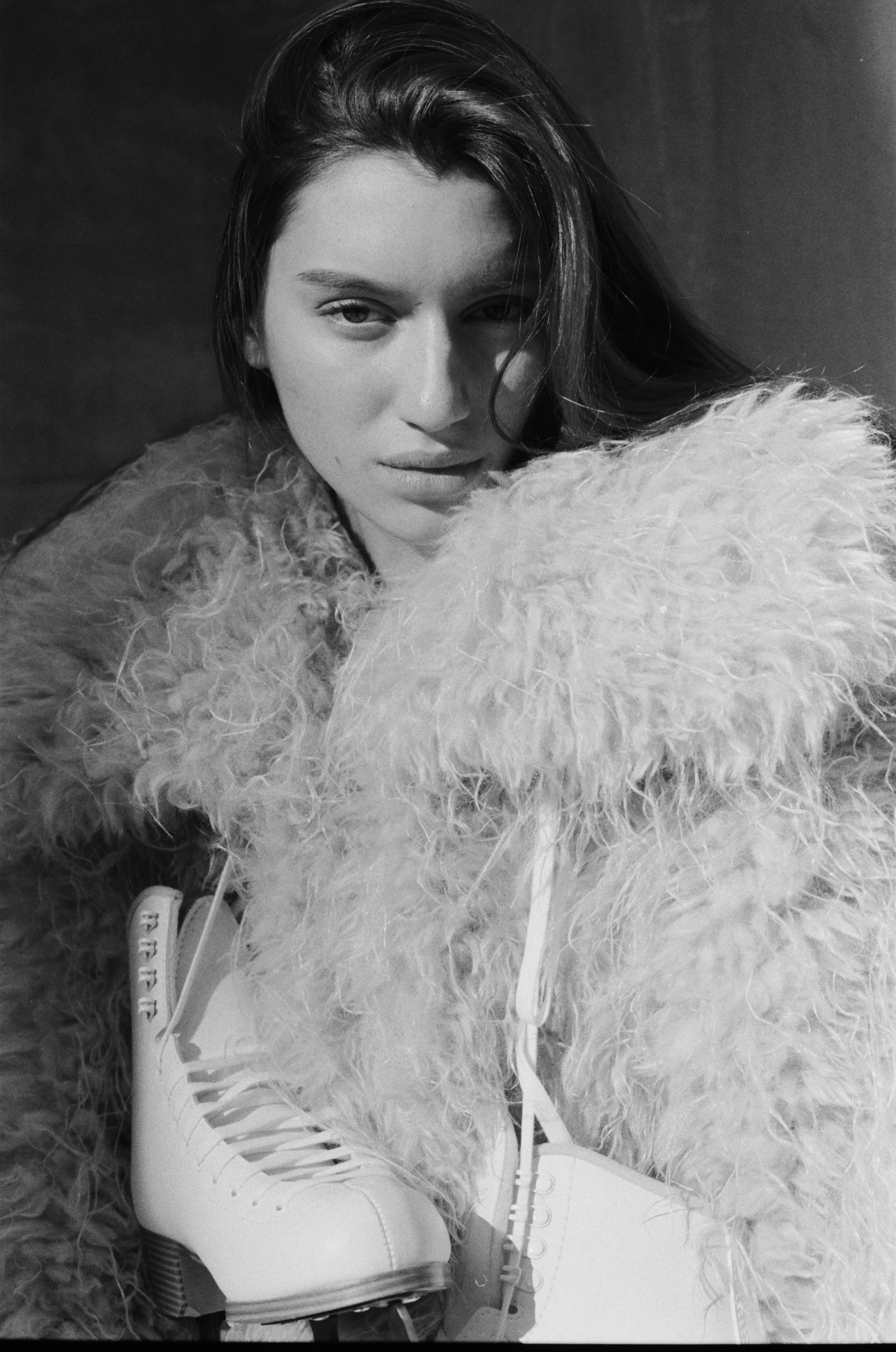 Black and white photo of a woman with long hair, wearing a fluffy coat, and ice skates.